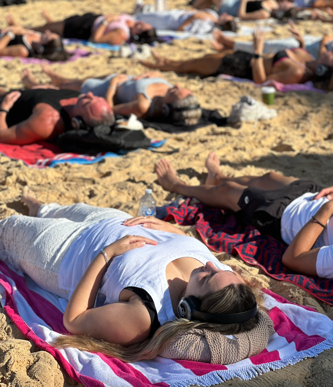 People doing breathwork beach towels on sandy beach, relaxing and enjoying the sunny weather, some wearing headphones.
