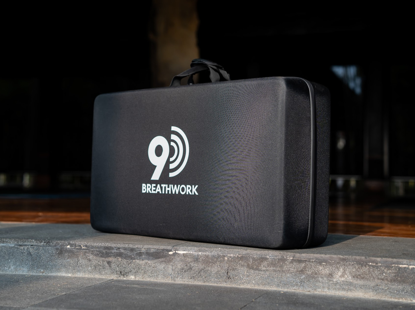 Black suitcase with white logo and the words '9D BREATHWORK' on the front, placed on a concrete surface outside.