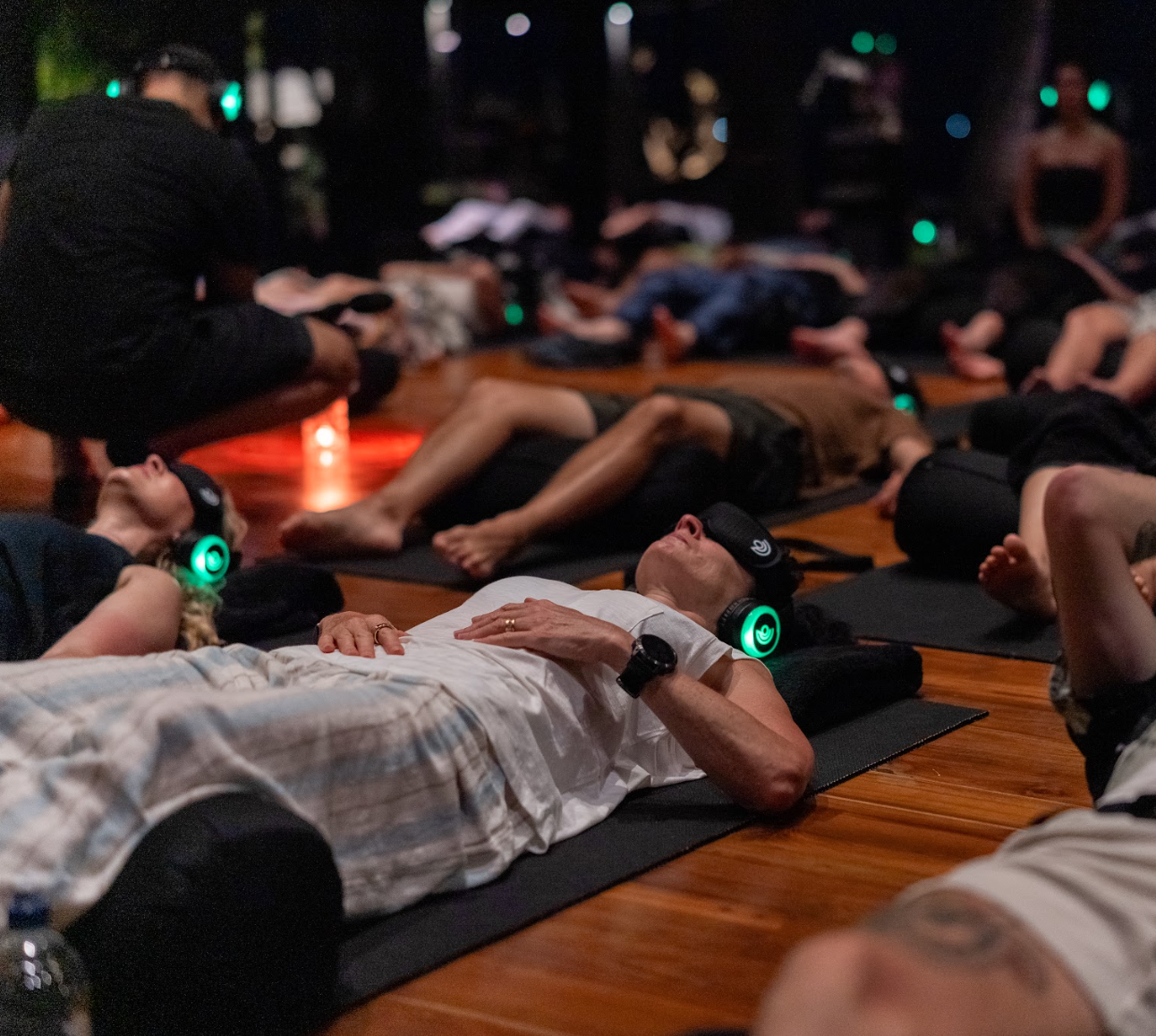People participating in a group meditation session in a dimly lit room, lying on yoga mats with eyes covered by headbands and wearing headphones, surrounded by others in a similar position.