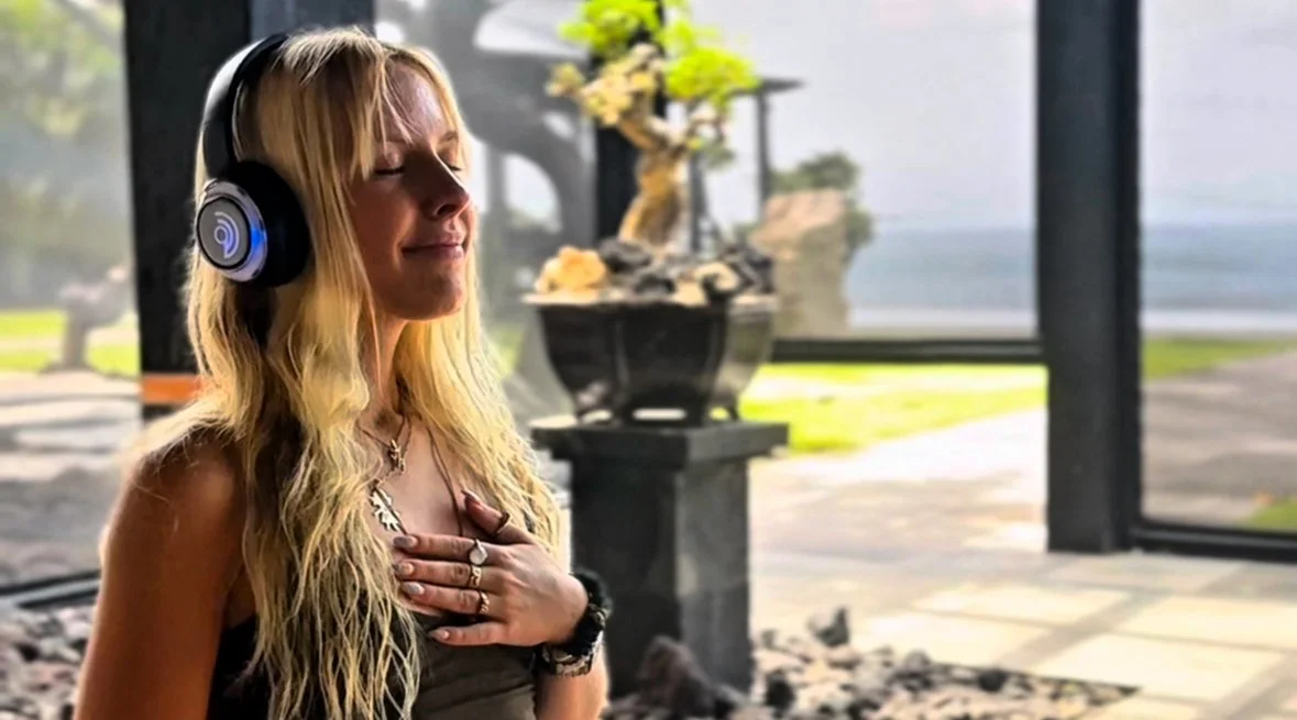 A person standing near the water with arms raised above their head, facing the sea on a cloudy day.