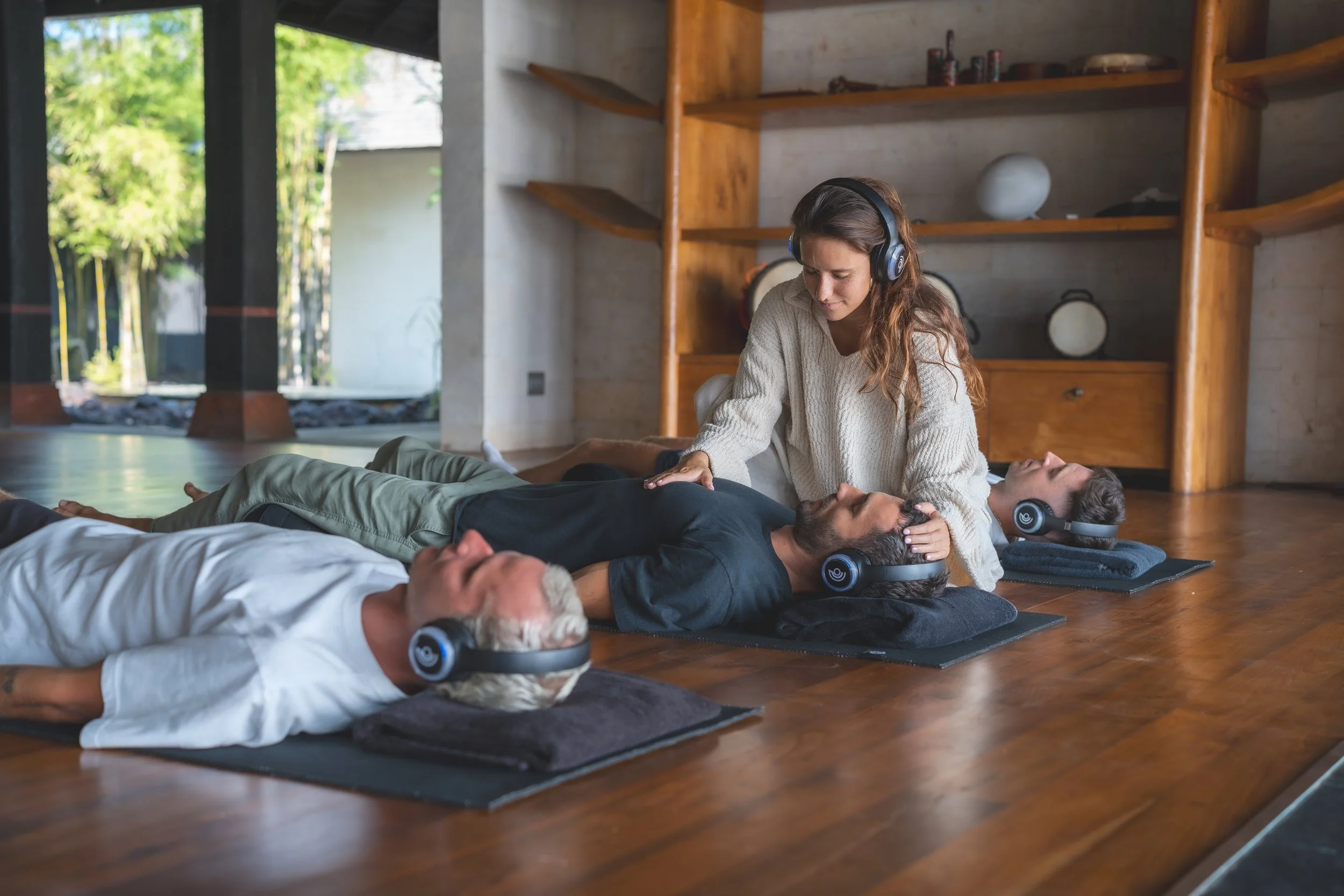 Two people relaxing outdoors on yoga mats by a pool, wearing eye masks and headphones, sunbathing on a sunny day.