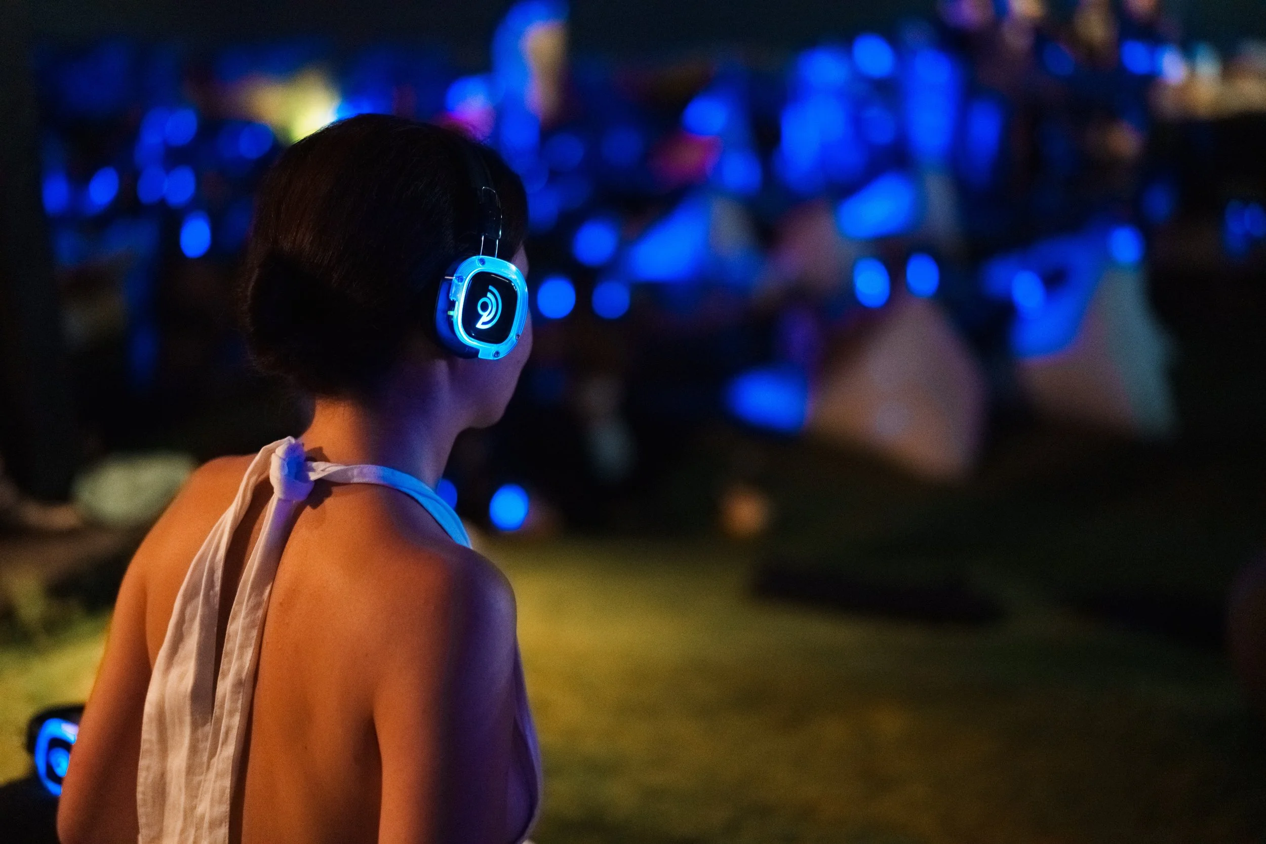 A woman with short dark hair wearing illuminated blue headphones, standing outdoors at night with a crowd and tents illuminated in blue in the background.
