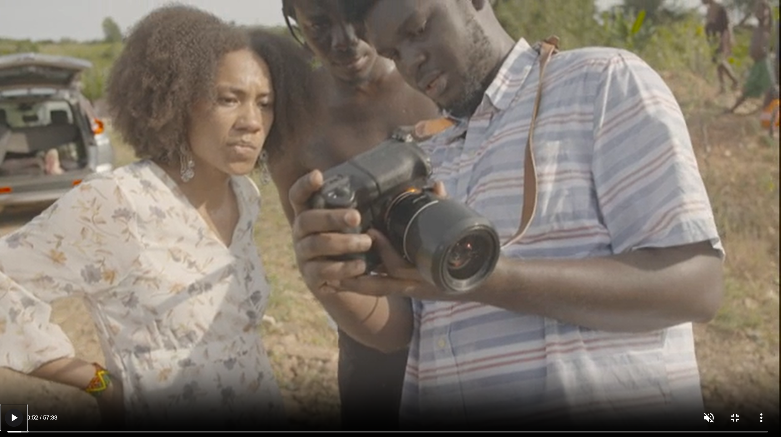 Three people examining a camera outdoors during daytime, with a background of vehicles and trees.