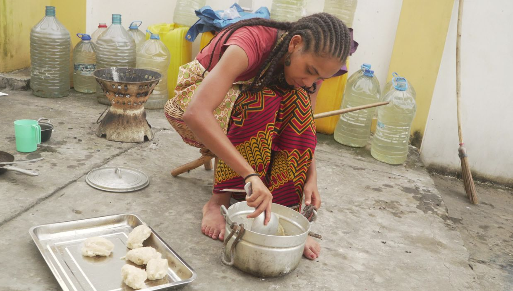 A woman with long braided hair, sitting on the ground. She is barefoot and surrounded by containers of water and other household items.