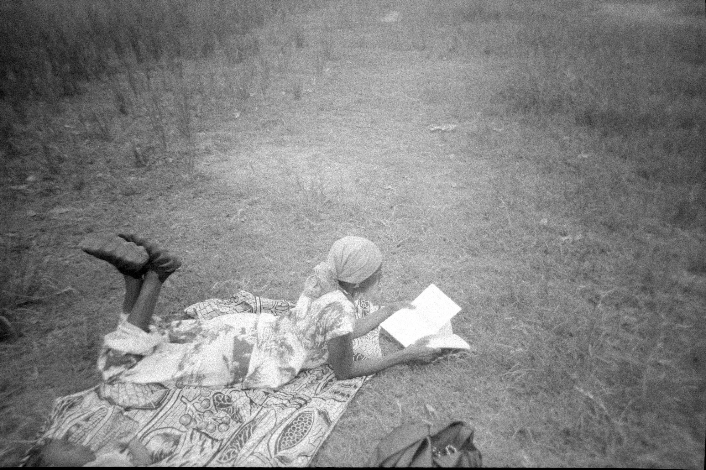 Woman lying on patterned cloth on grass, outdoors, with hands holding a book, wearing a headscarf.
