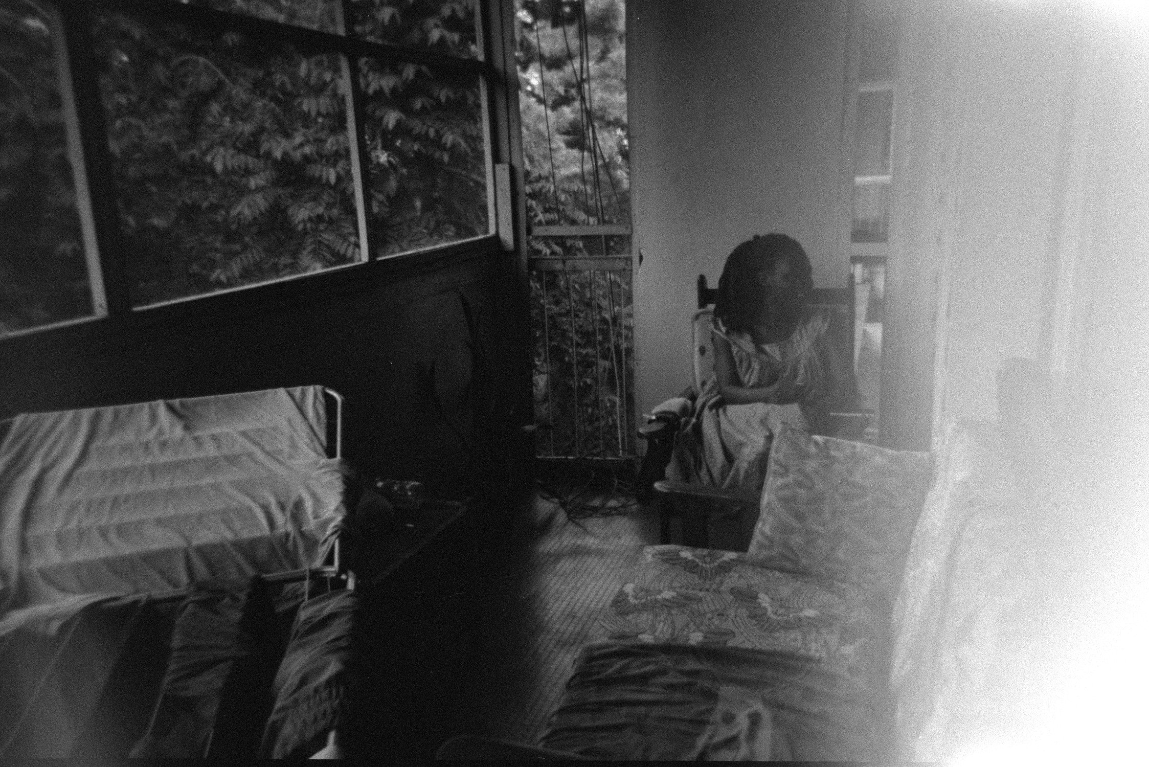A black and white photo of an indoor room with large windows showing trees outside. There are beds, a person sitting on a chair, and assorted furniture and belongings scattered around.