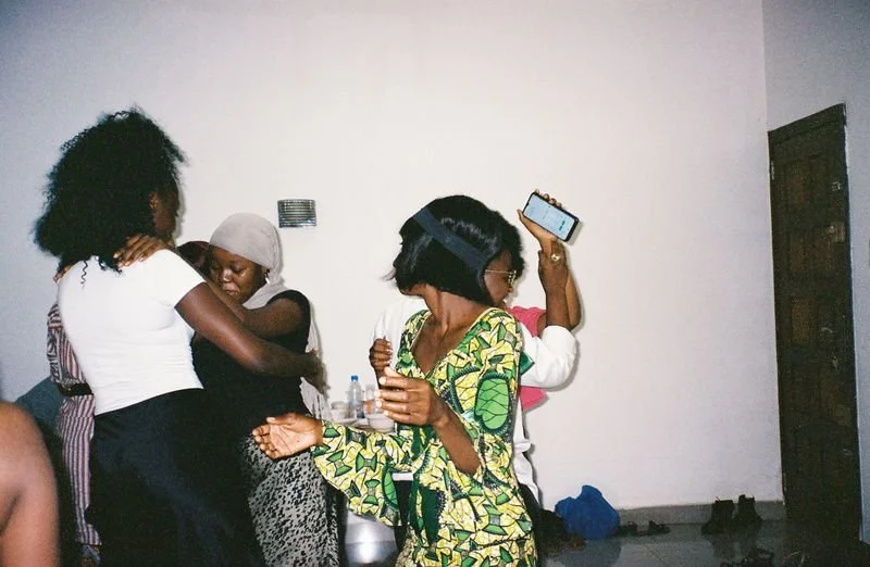 Four women are gathered in a room, with one woman holding a smartphone and others hugging or engaging in conversation.