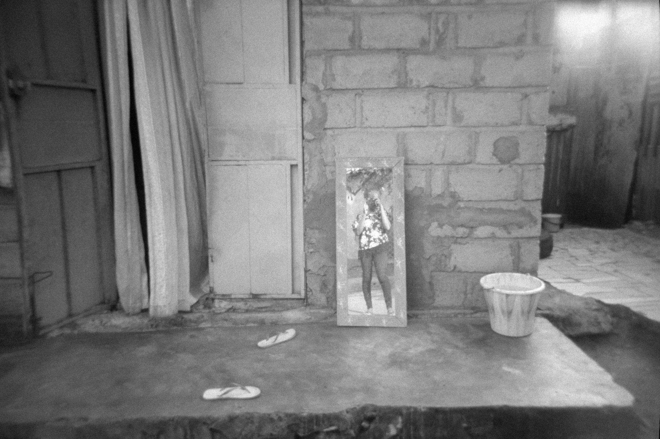 A black and white photo of a room with brick walls and a concrete floor. There is a full-length mirror reflecting a person taking the photo, standing in front of the mirror.