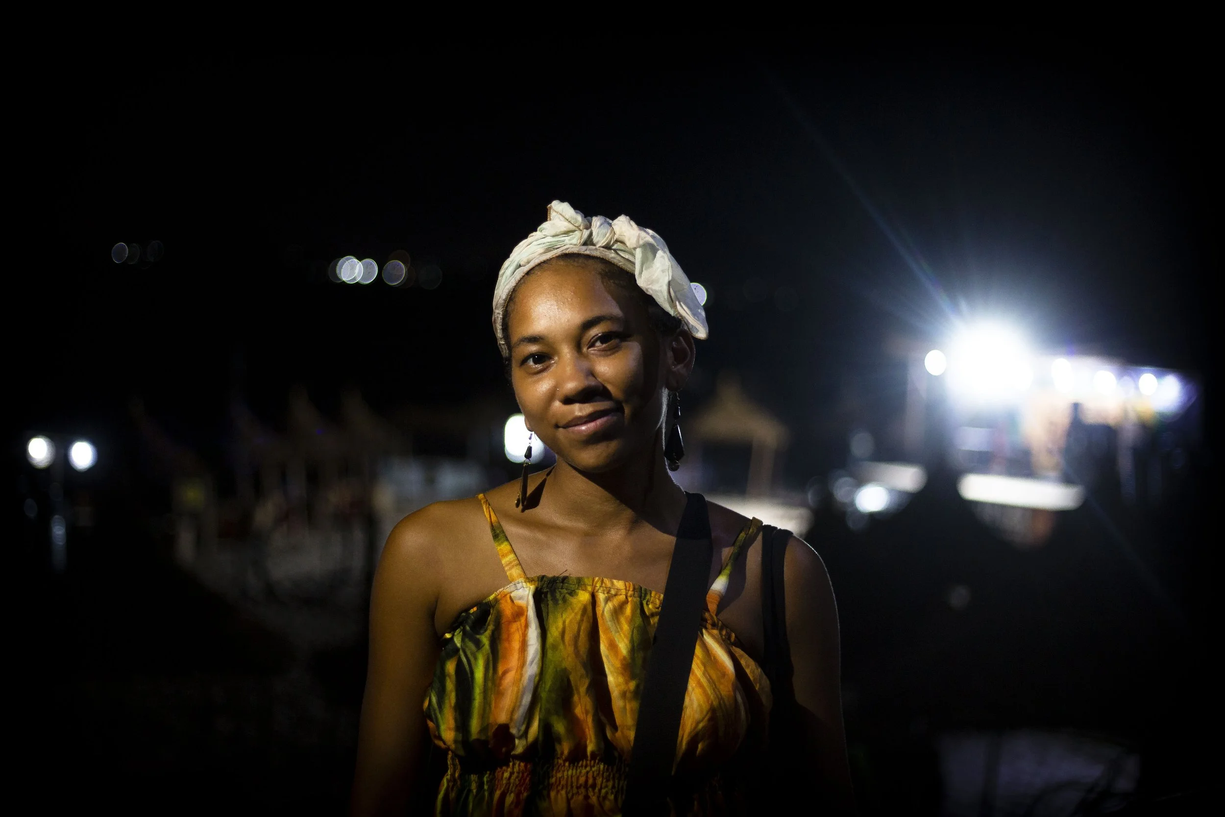A young woman with a light headscarf, wearing a colorful dress, stands outdoors at night with bright lights in the background.