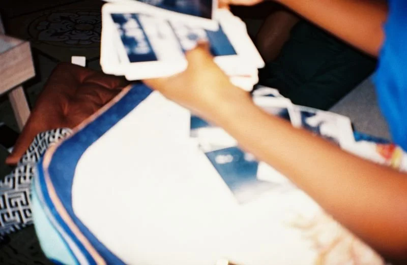 A person holding a fan of photographs or postcards while sitting at a table, with various items including a leather chair and patterned tablecloth visible around.