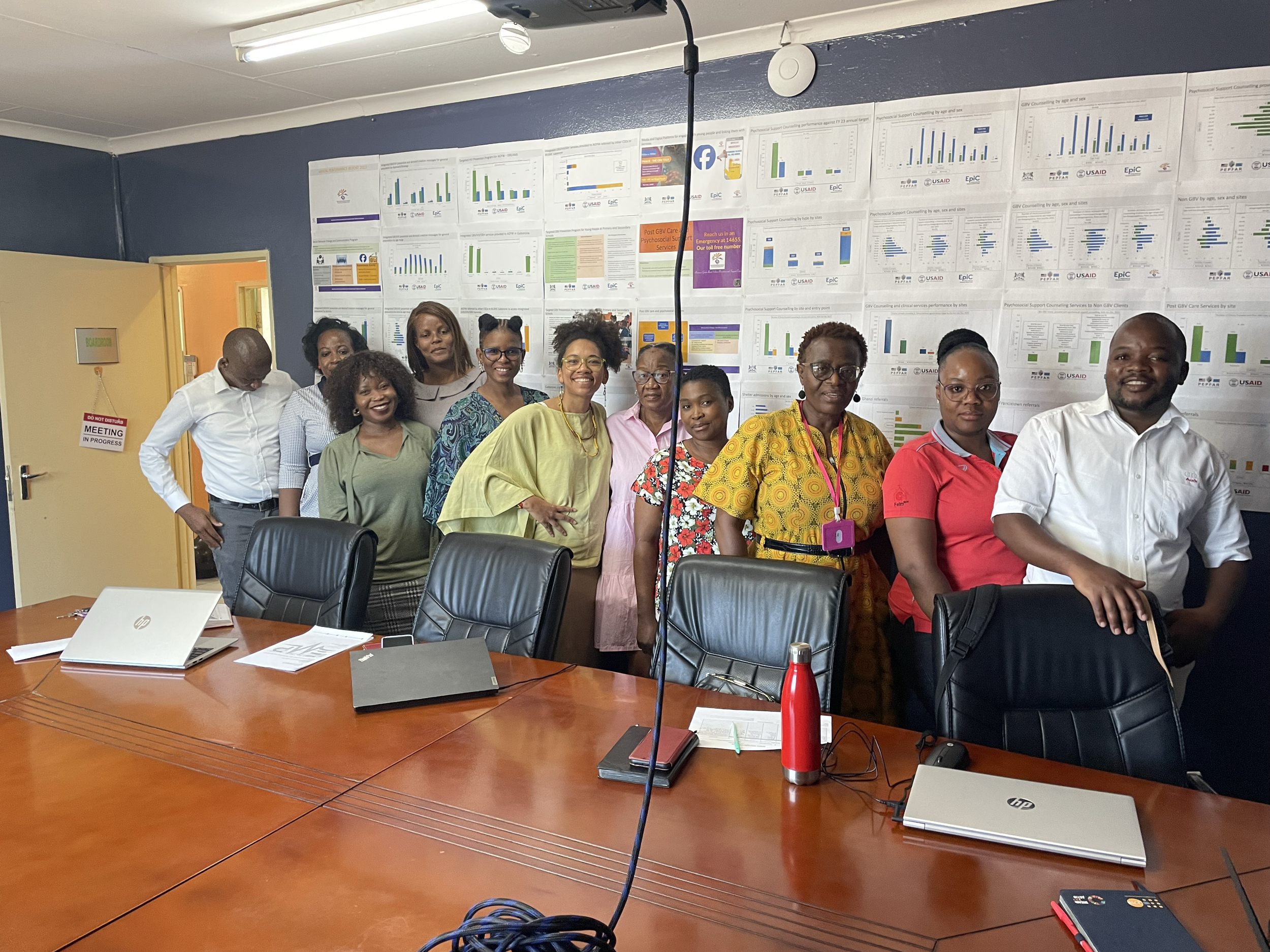 Group of twelve people standing behind a conference table in a room with charts and graphs on the wall.