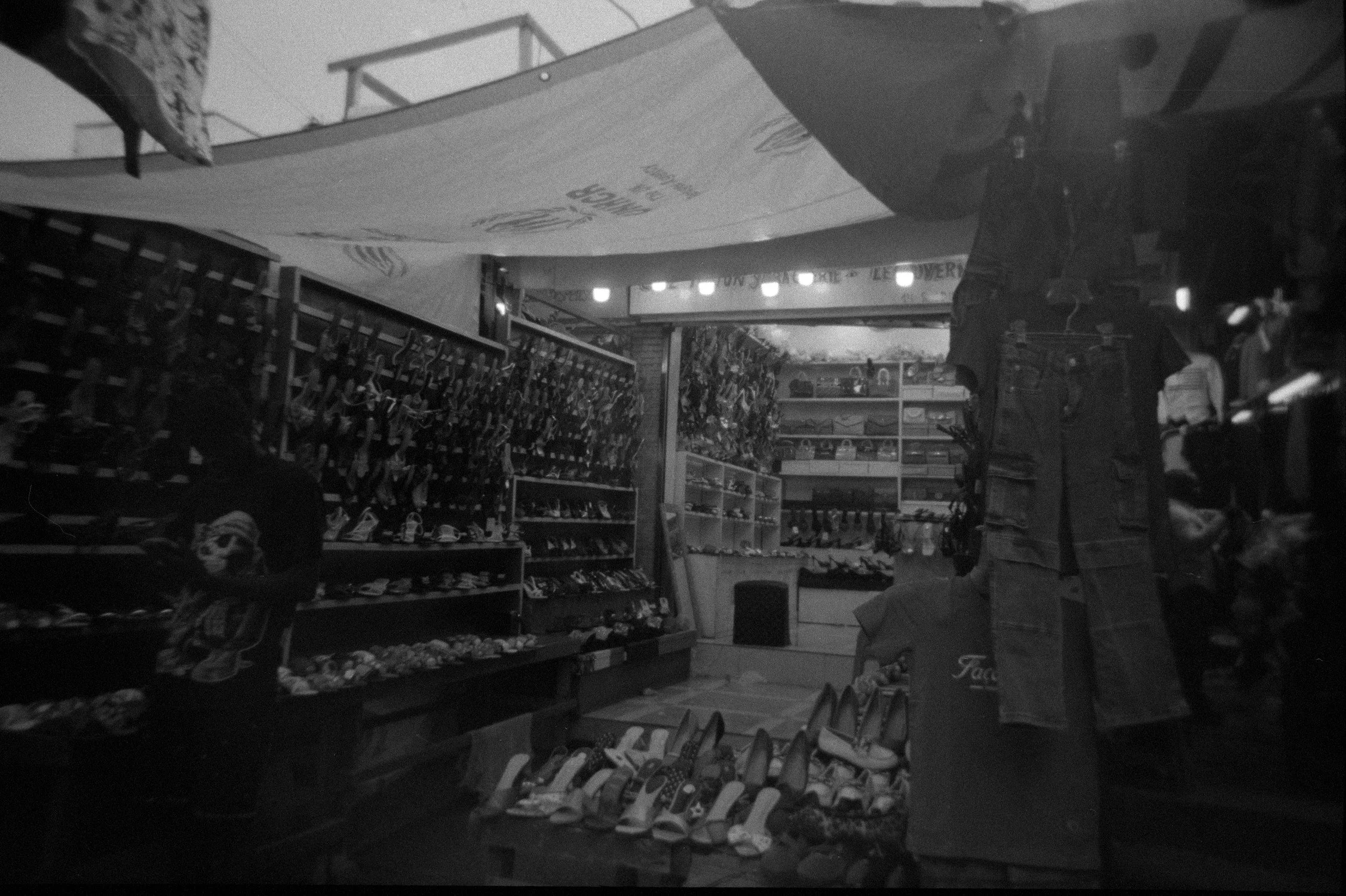 Black and white photo of a store aisle with shelves filled with shoes and accessories, with a person browsing on the right side.