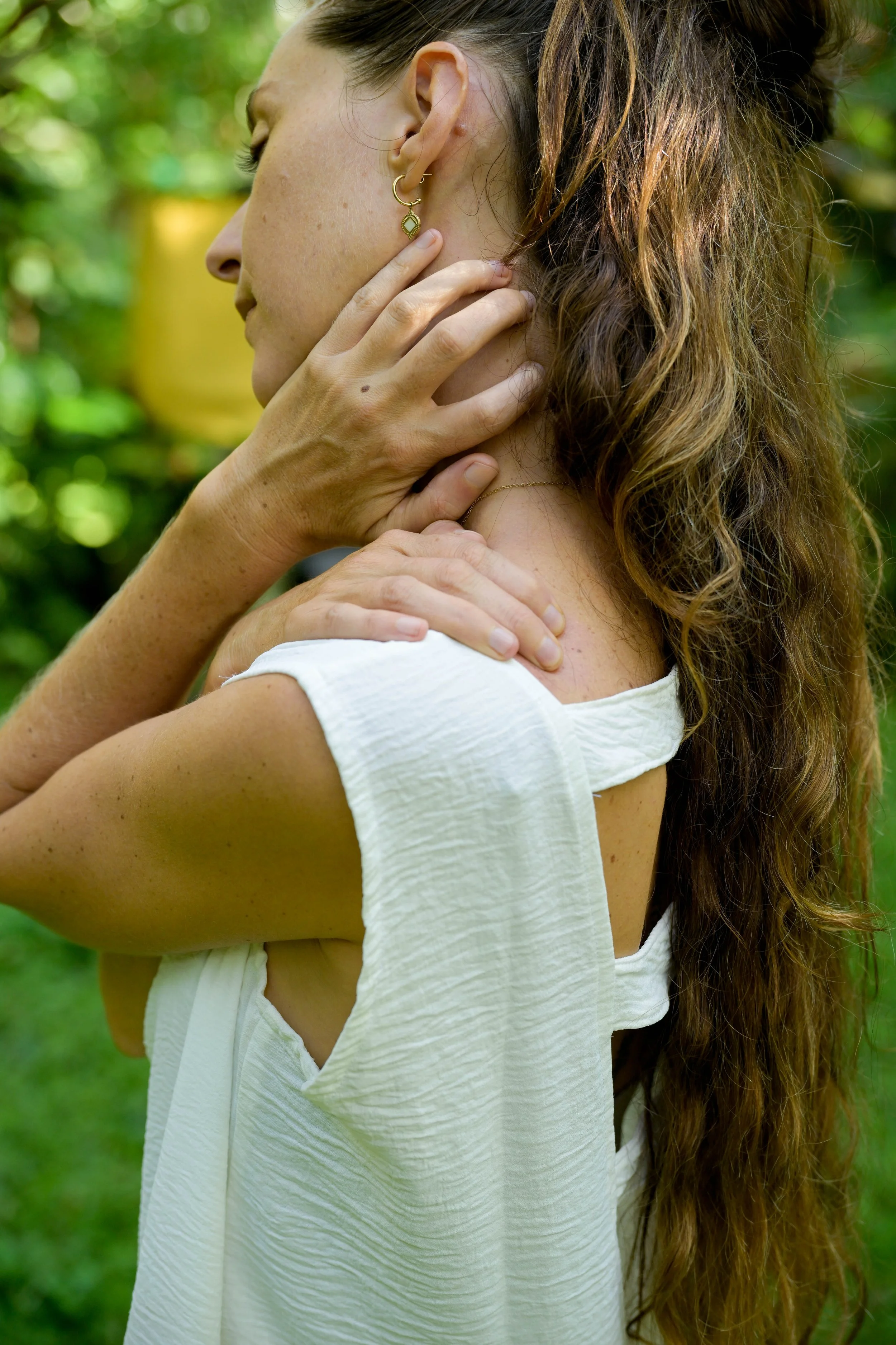 A woman with long curly hair wearing a white top and gold jewelry is gently holding her neck and shoulder, with a green outdoor background.