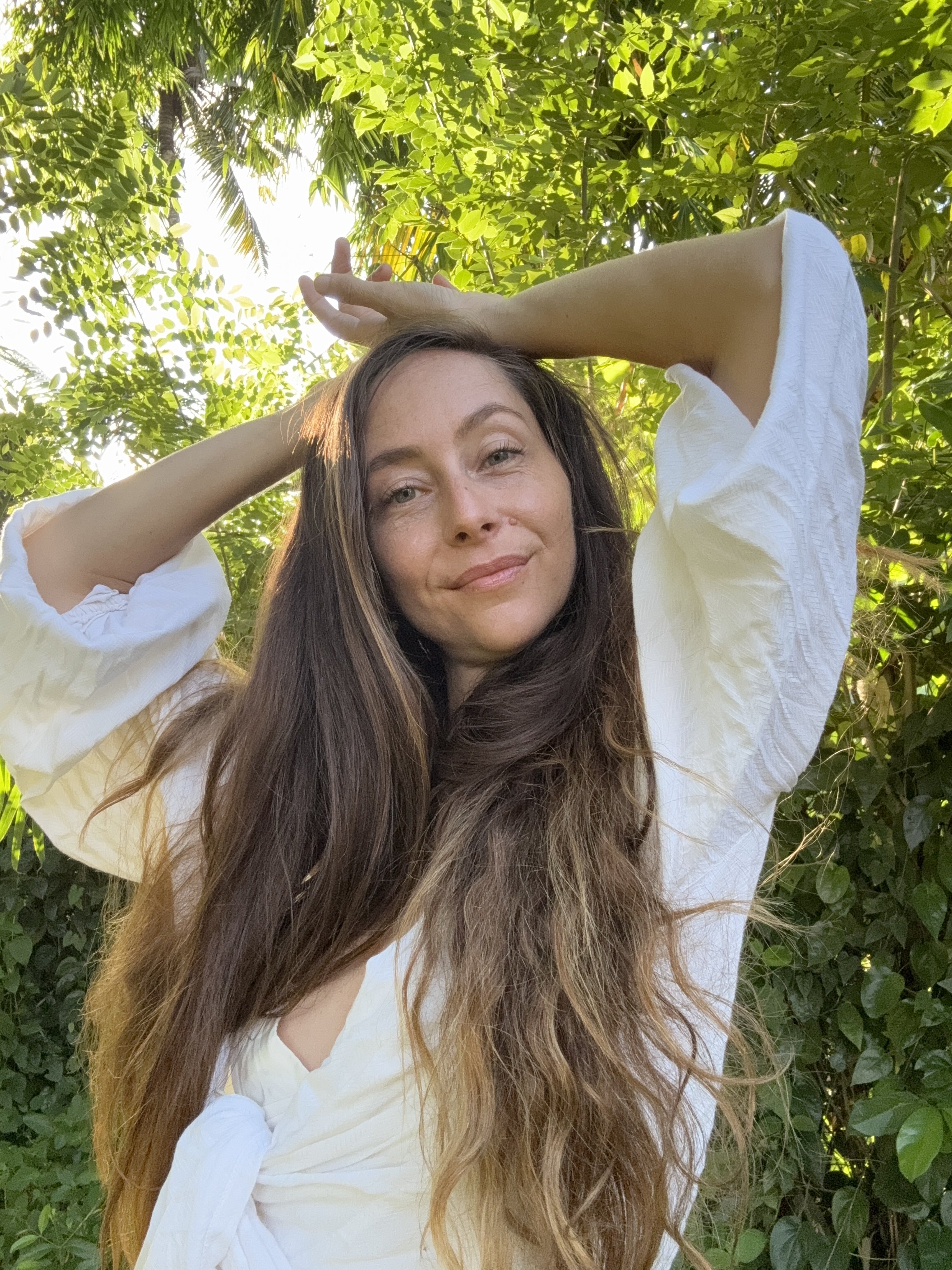 A woman with long brown hair and light skin smiling outdoors among green foliage, wearing a white blouse with her arms raised above her head, backlit by sunlight.