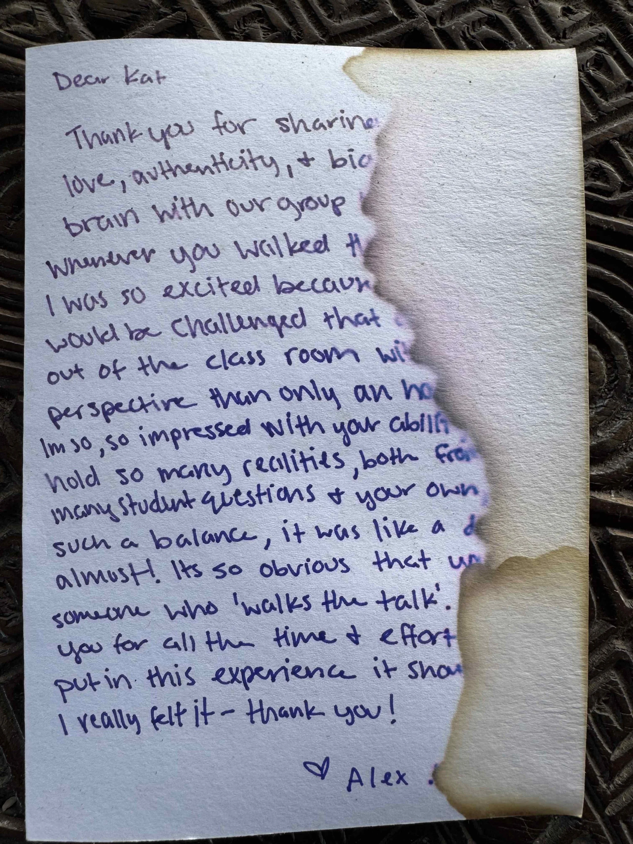 Handwritten thank you note on a piece of paper with burnt edges, placed on a dark textured surface.