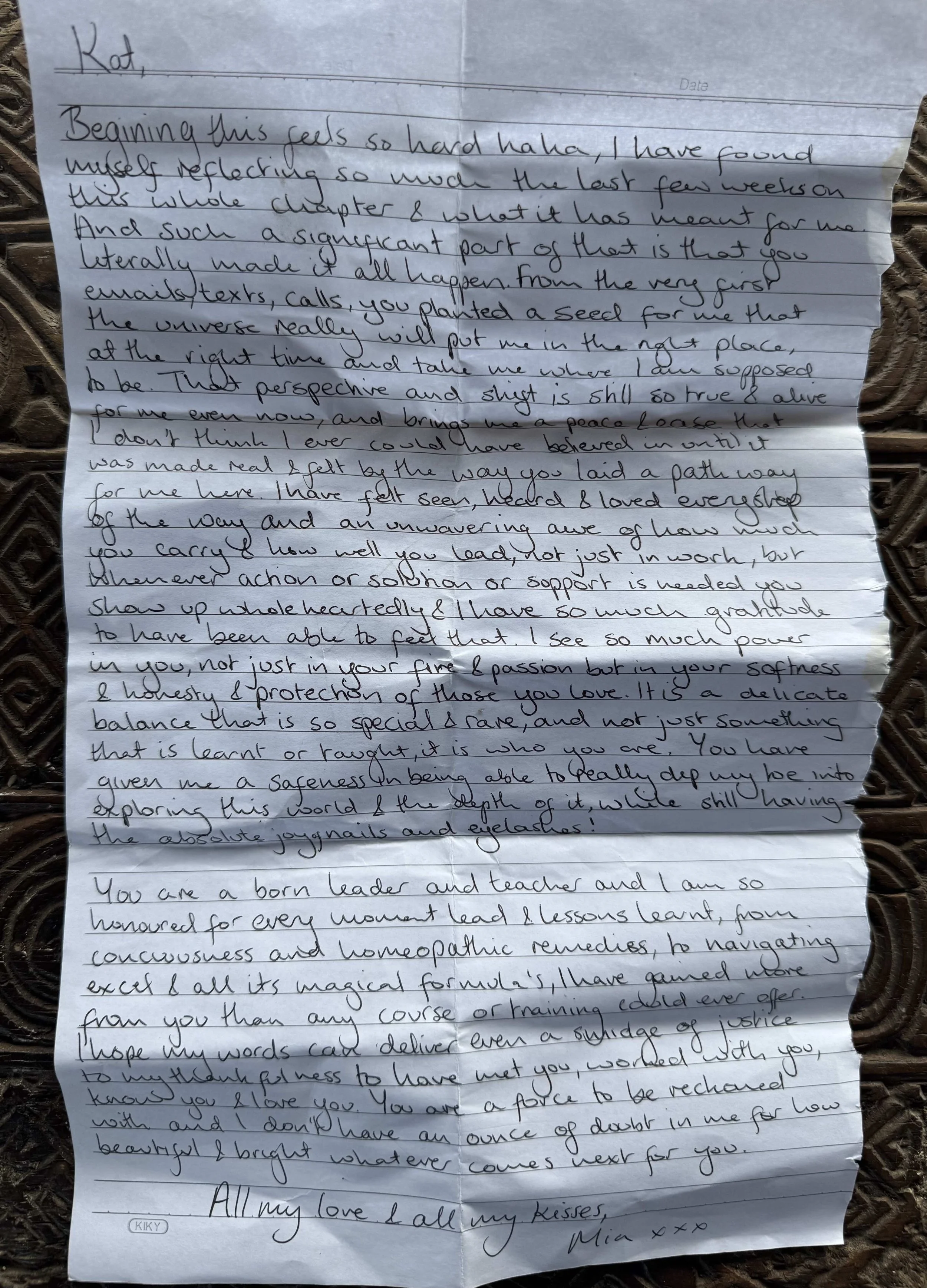 A handwritten letter on lined paper with some creases, placed on a woven surface. The letter is addressed to Kat and signed by Mia.