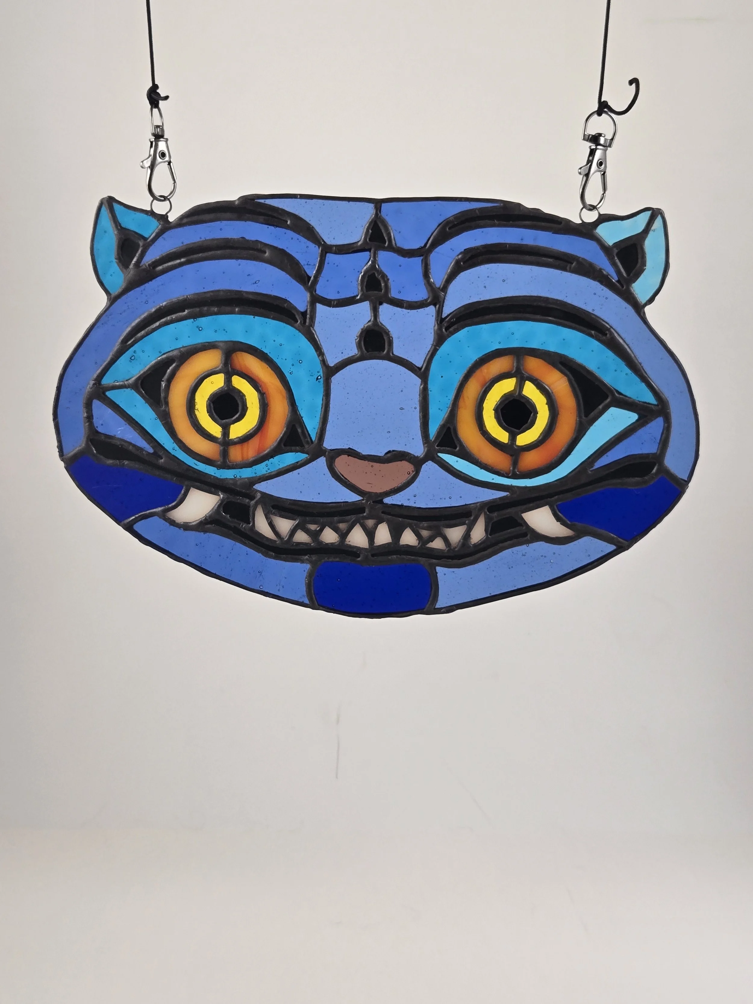 Colorful stained glass suncatcher in the shape of a cat's face with large yellow eyes and a pink nose, hanging from two wires with hooks.