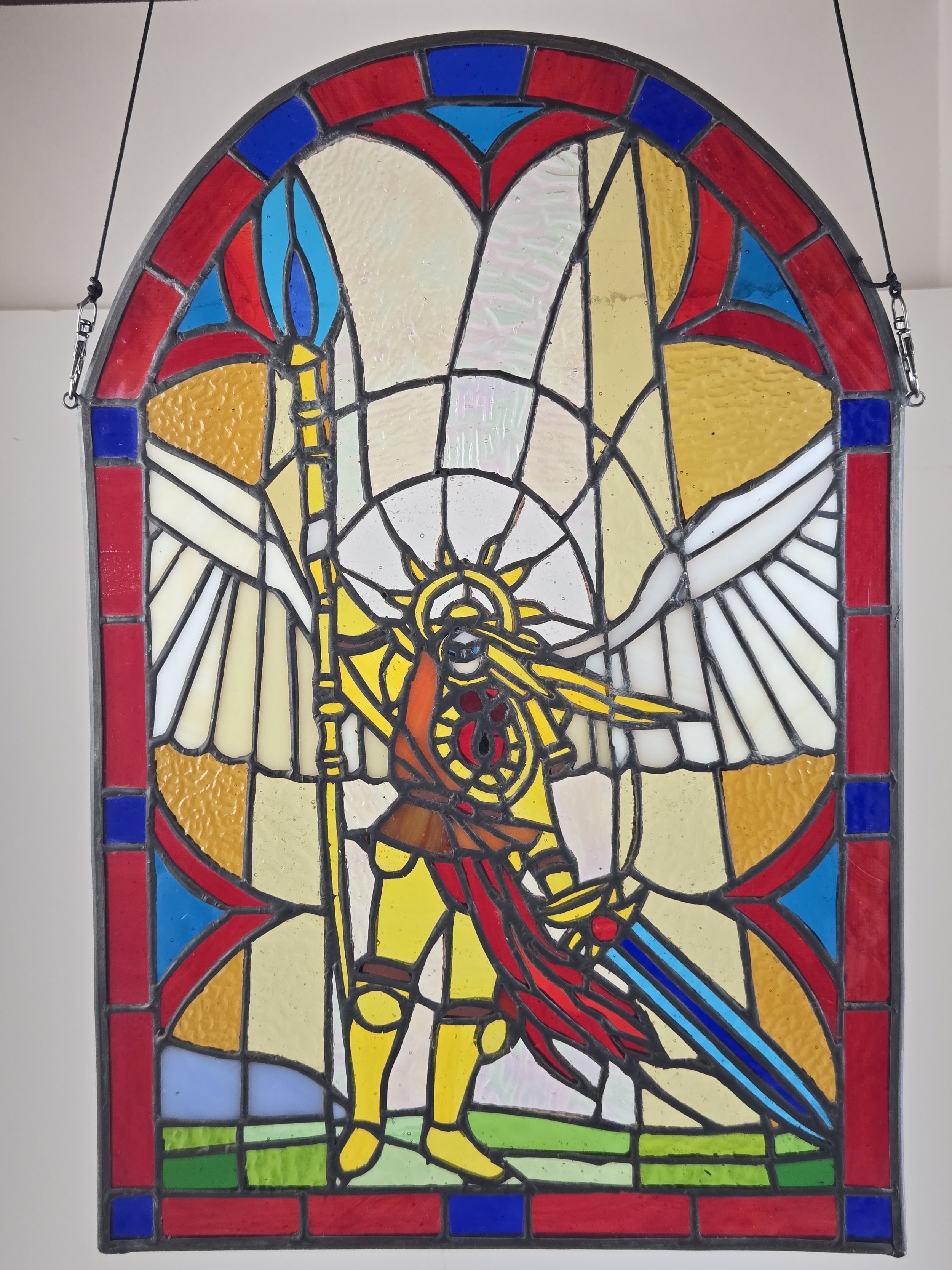 Colorful stained glass window depicting an angel with wings, holding a sword and a shield, surrounded by a red and blue border.