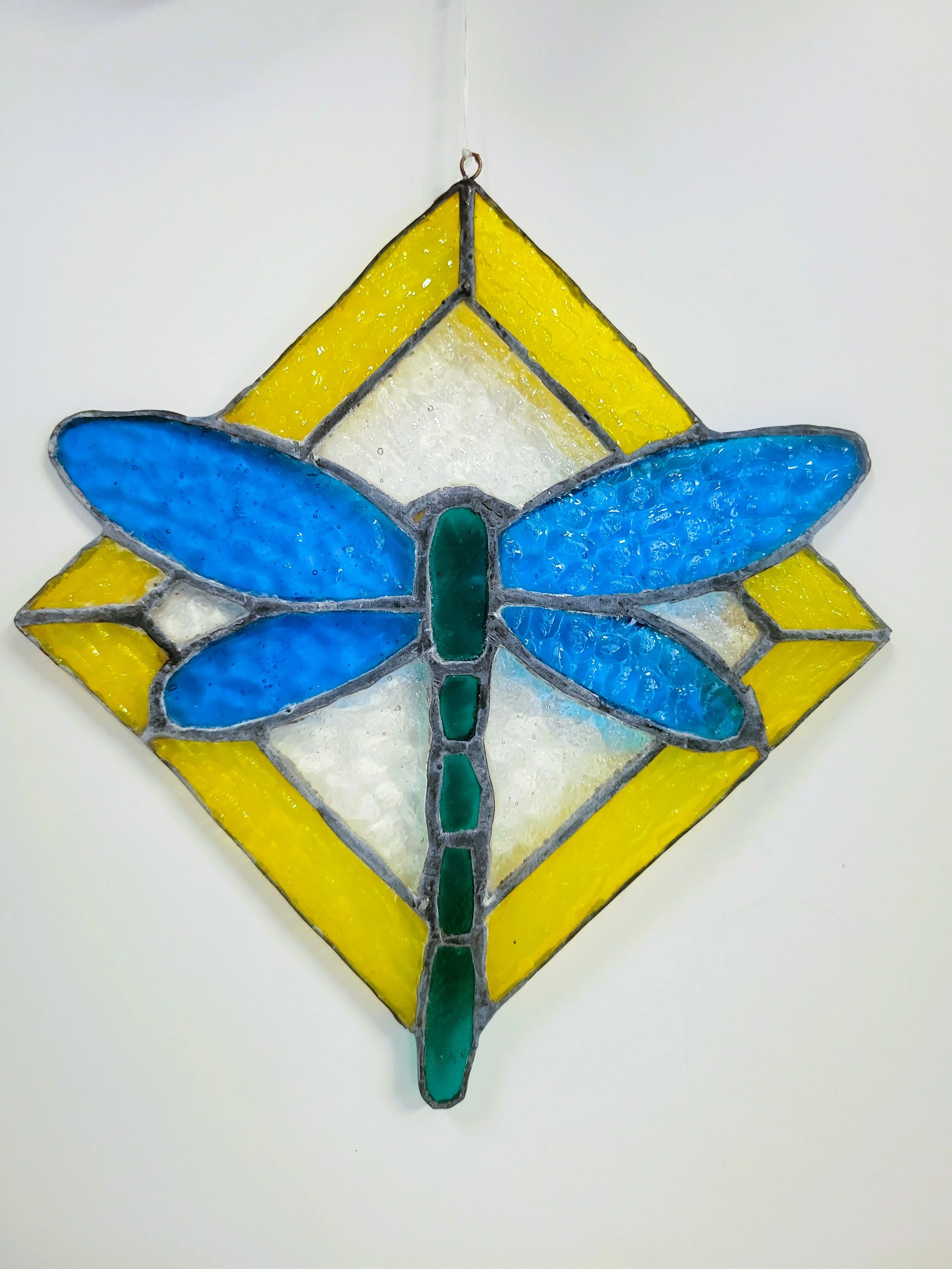A stained glass suncatcher with a blue dragonfly design, set against a yellow and white geometric background.