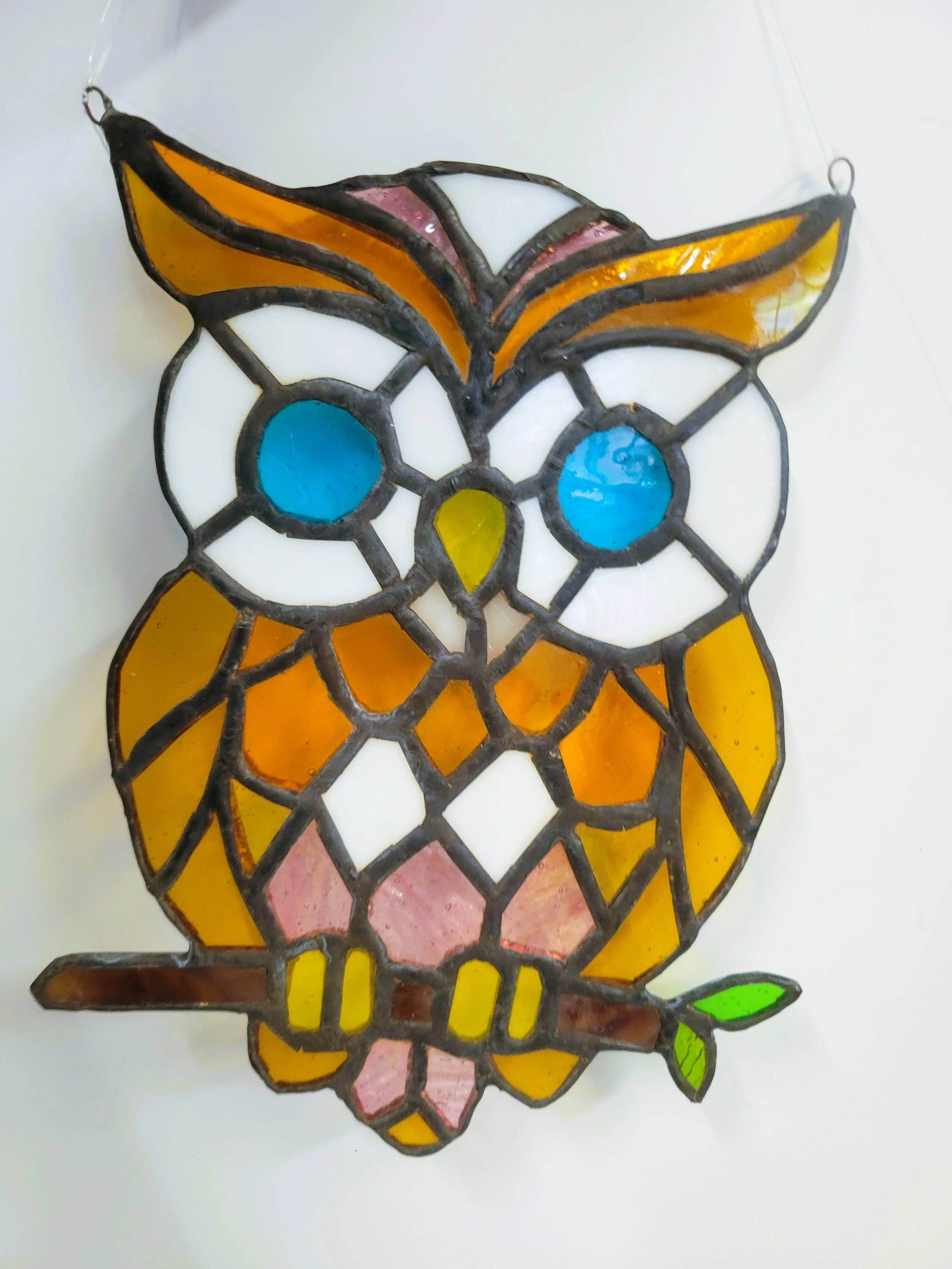 Colorful stained glass owl decoration with orange, yellow, pink, blue, and green glass pieces, hanging on a white wall.