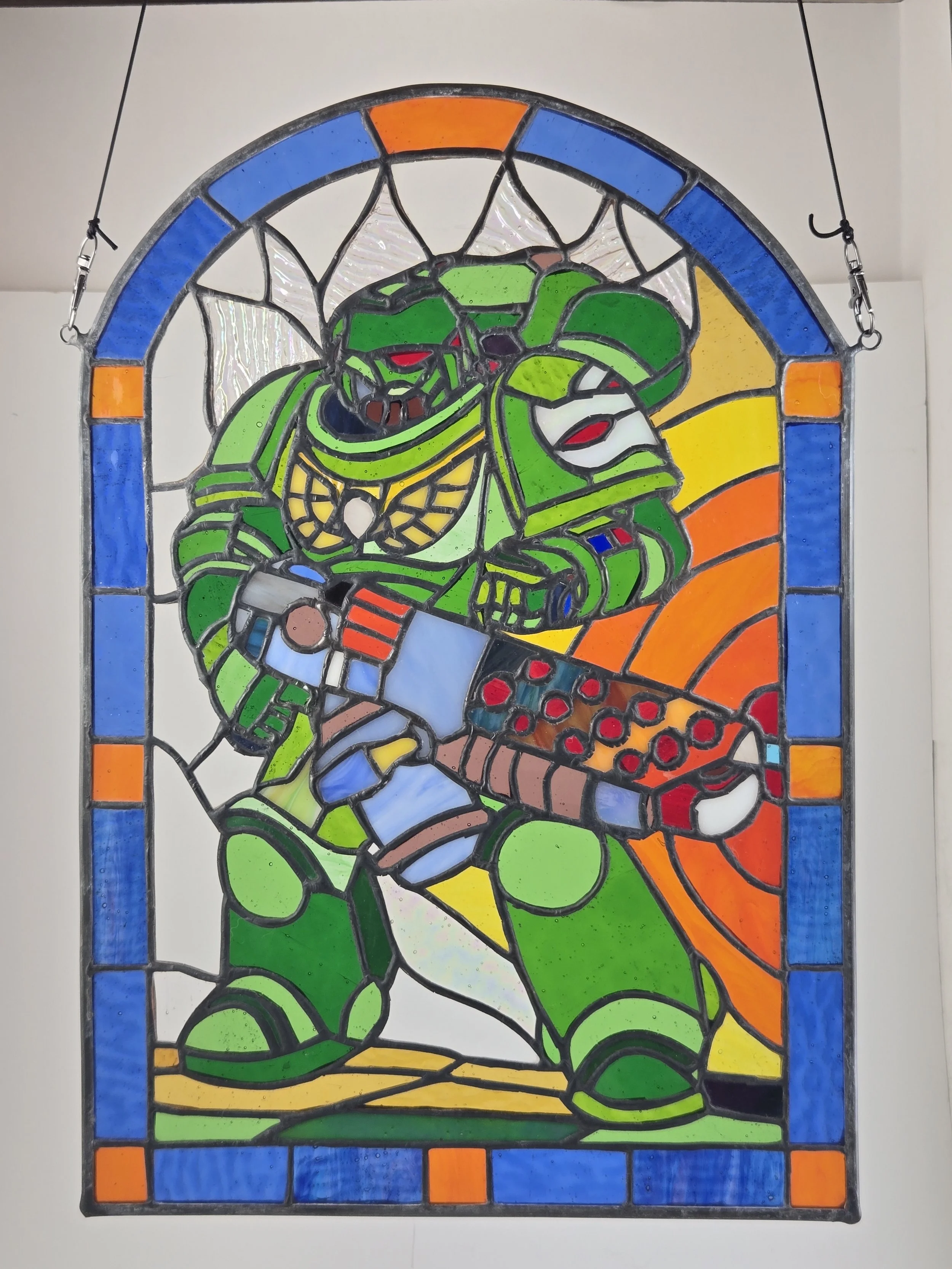 A stained glass piece of a futuristic soldier in green armor holding a weapon, with a colorful background and a border of blue and orange.