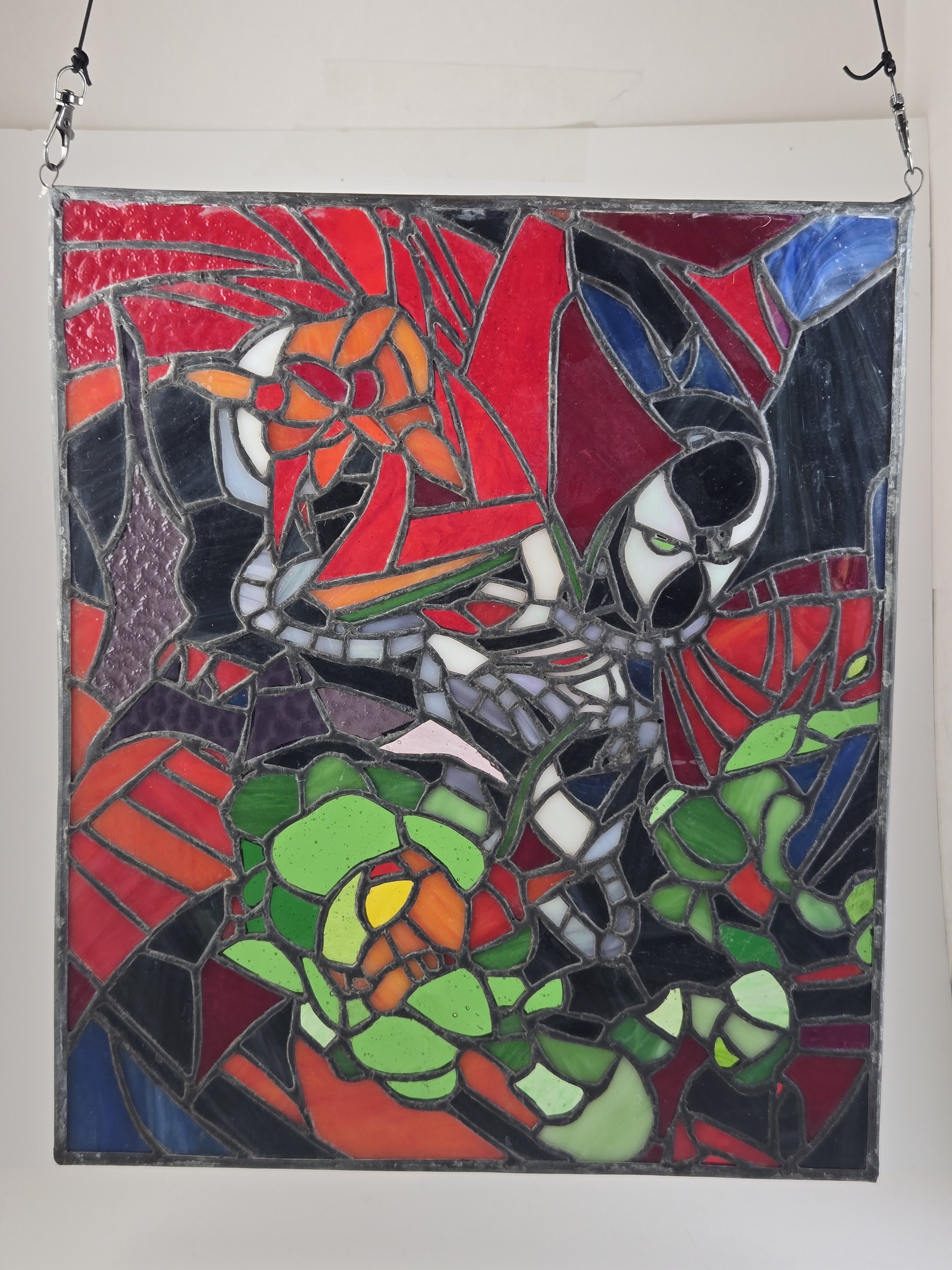 Colorful stained glass window depicting Spiderman in red and black, with green and black eyes, holding a green figure with an orange face.
