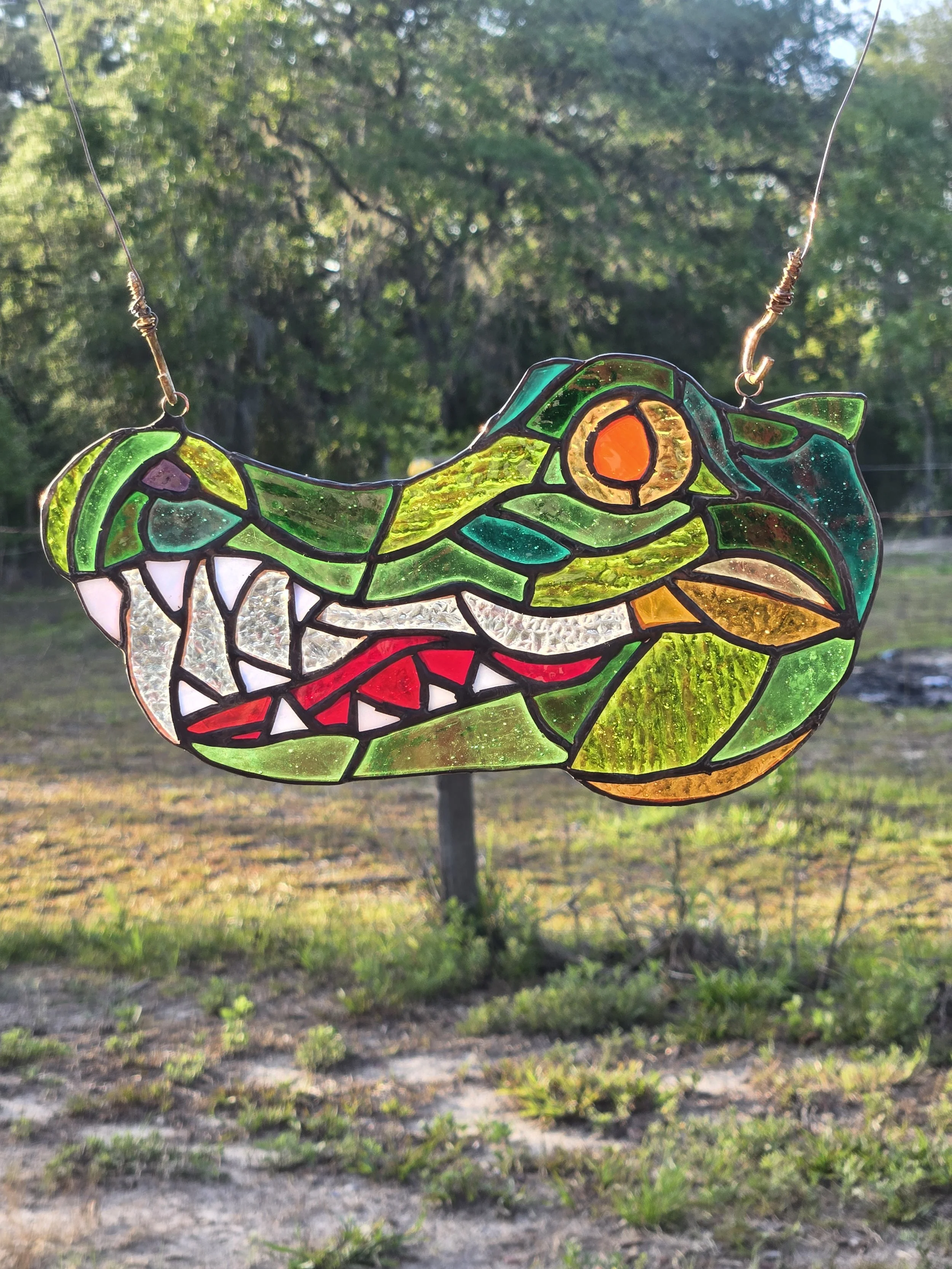 Colorful stained glass suncatcher depicting a alligator, hanging outdoors with trees and grass in the background.