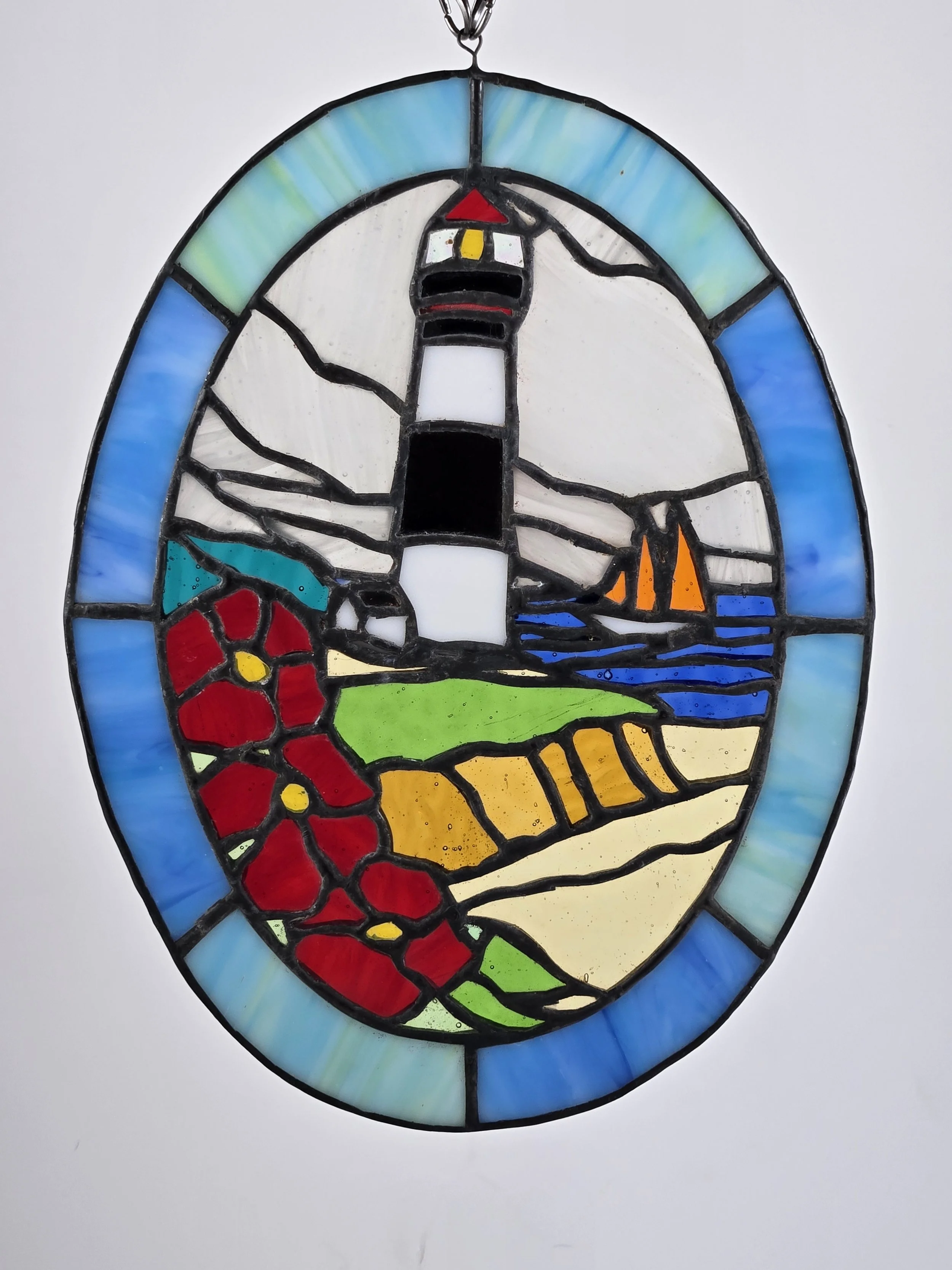 Stained glass piece of a lighthouse with sailboats, flowers, and landscape elements.