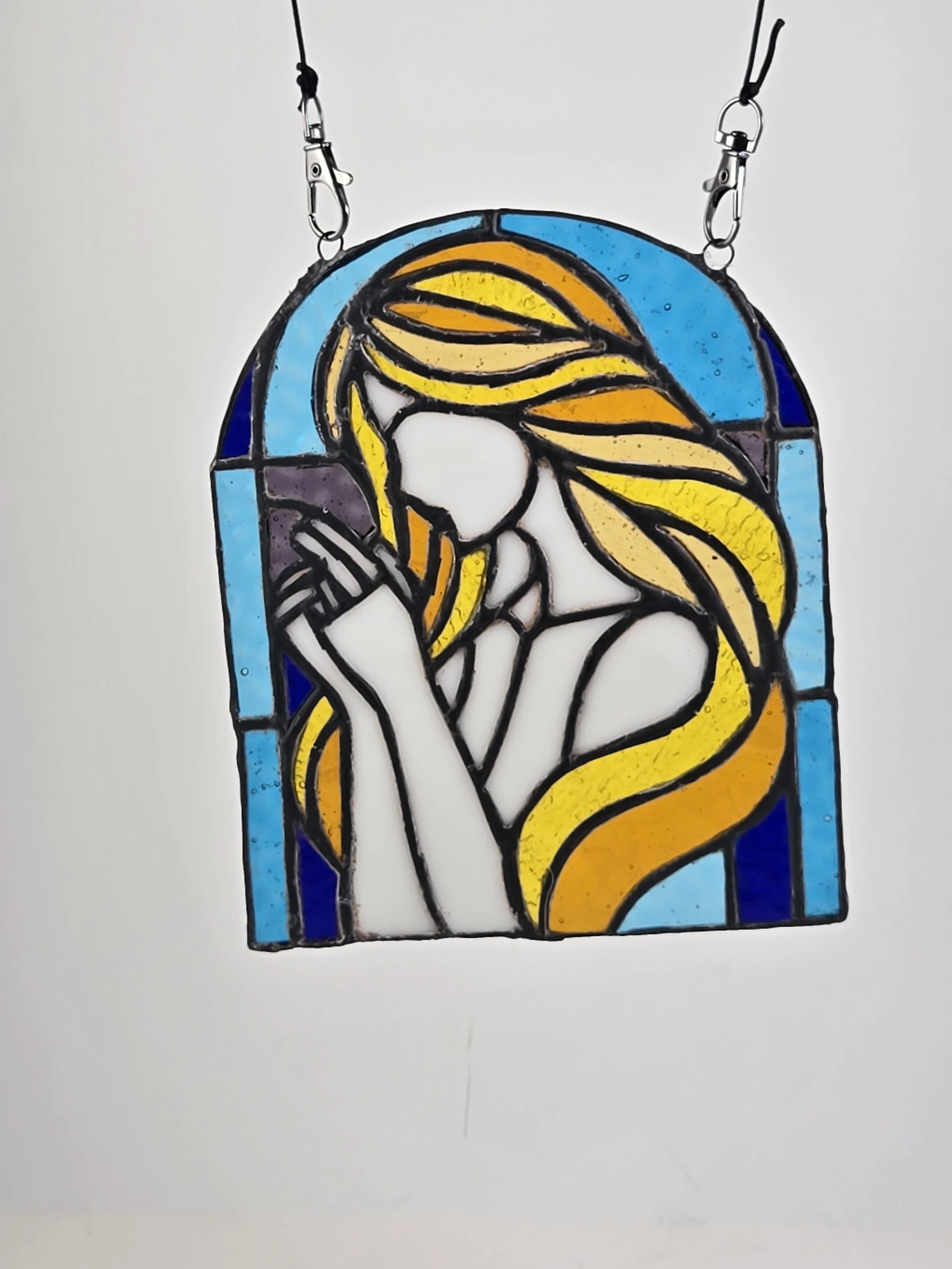 Stained glass artwork of a woman with long yellow hair, praying inside a rounded arch shape with blue background.