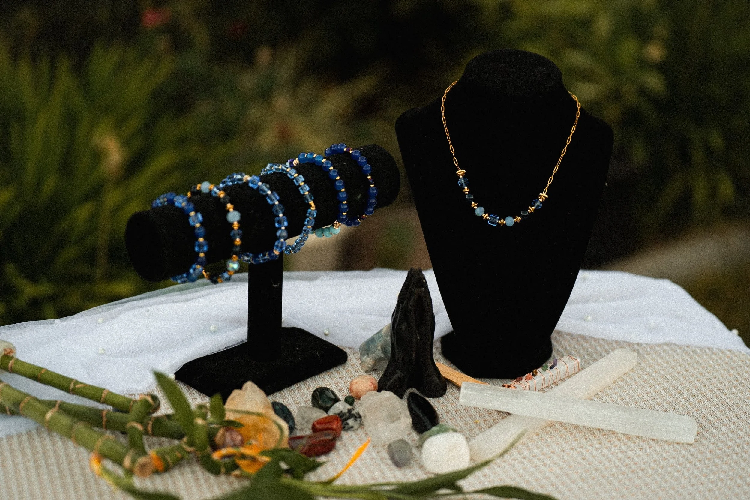 Jewelry display with a black necklace stand and a bracelet holder showcasing blue beaded jewelry, surrounded by crystals, stones, and a small white altar cloth, set outdoors with greenery in the background.