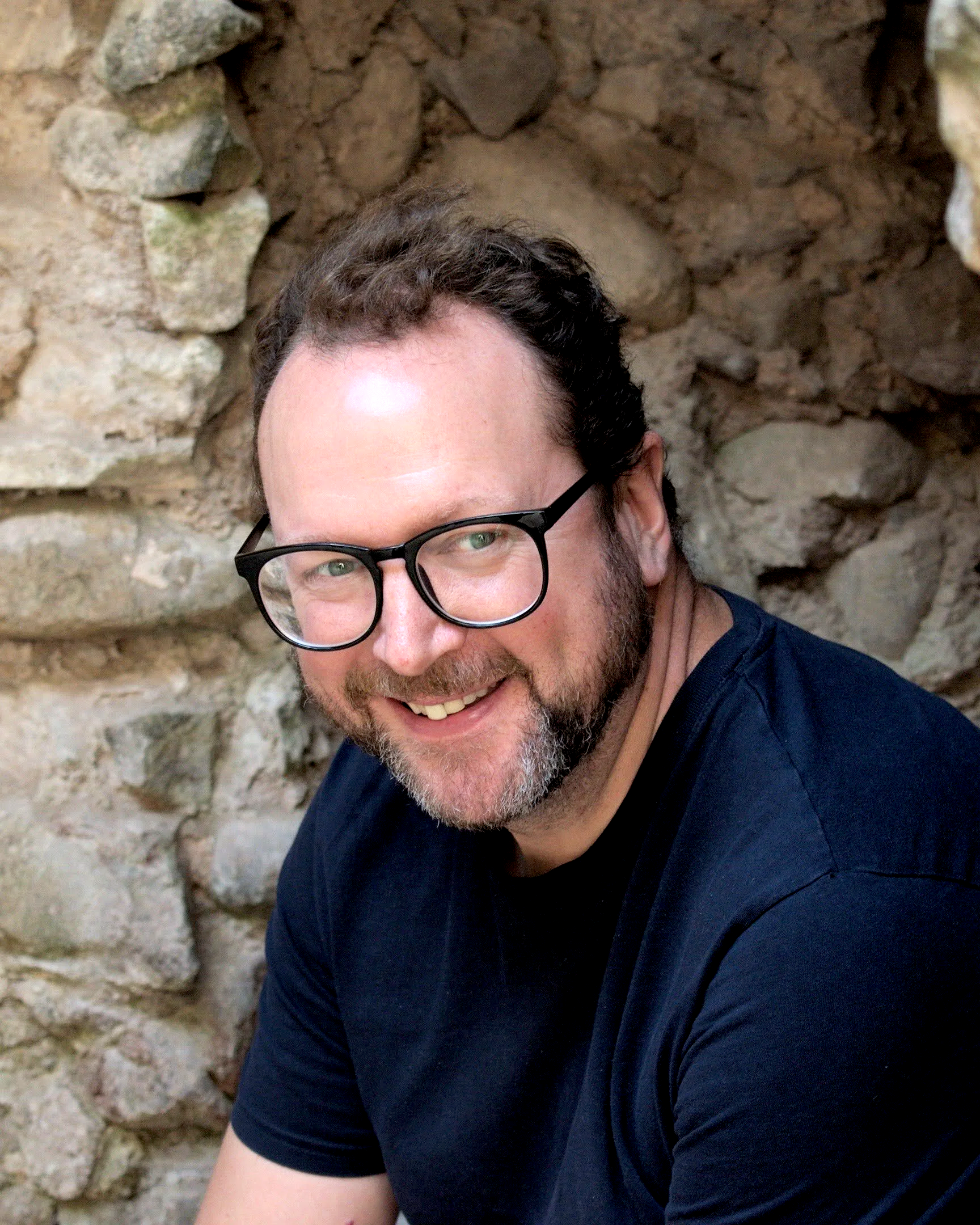 A man with glasses and a beard smiling at the camera, sitting outdoors near a stone wall.