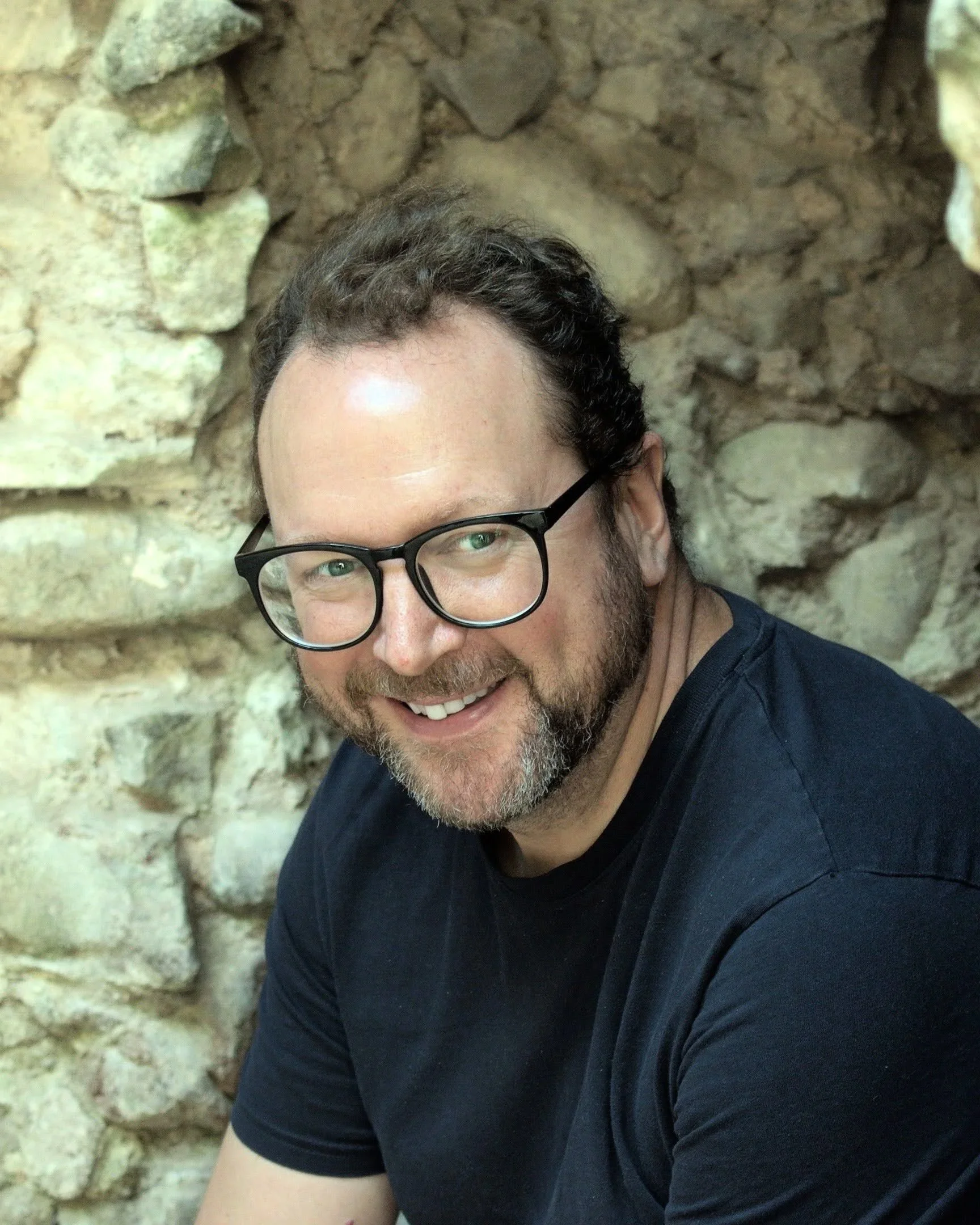 A man with glasses, a beard, and curly hair smiling, sitting in front of a stone wall.