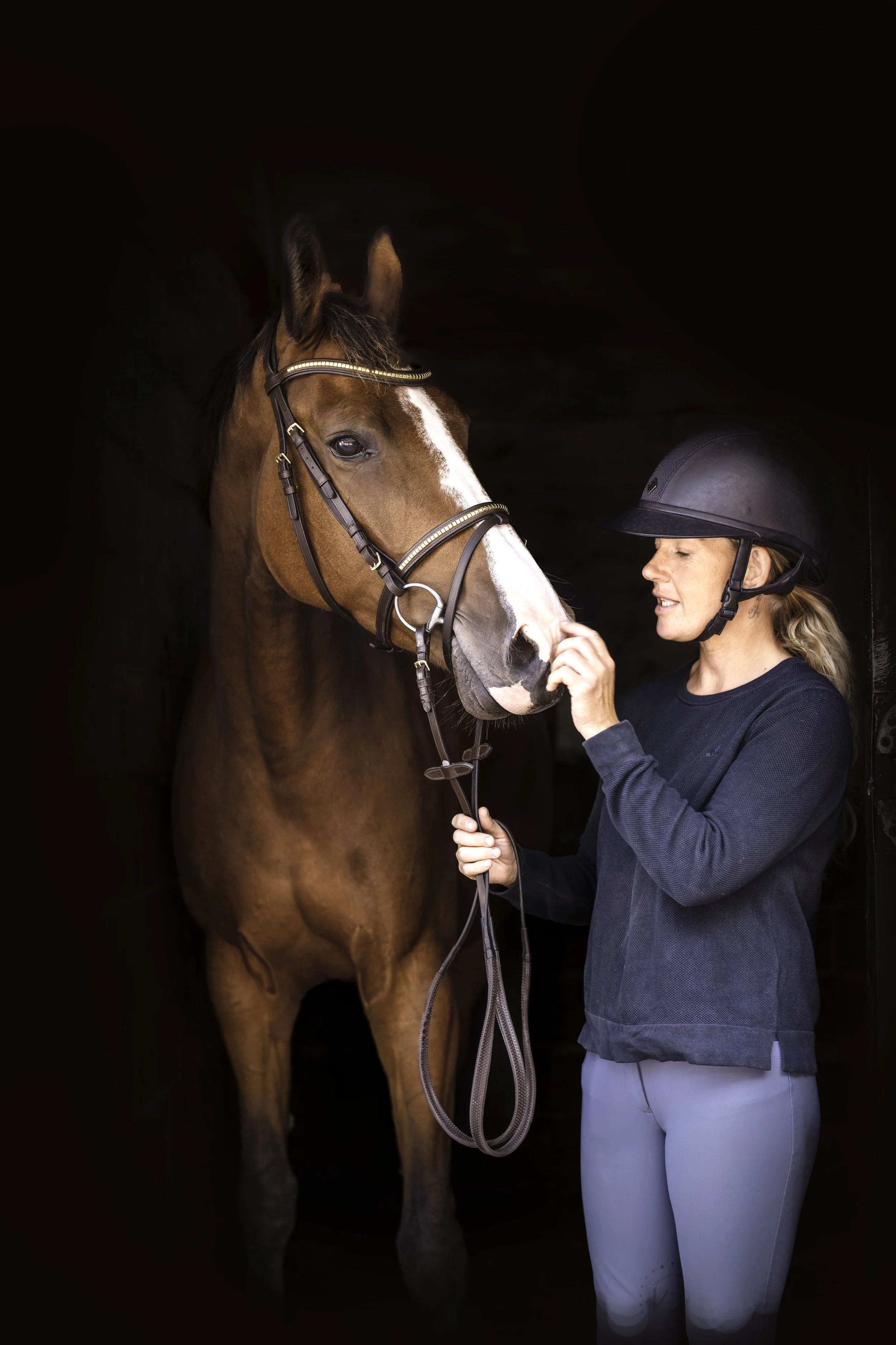 A woman in riding attire and a helmet gently touching the nose of a brown horse with a white blaze on its face inside a dark stable.