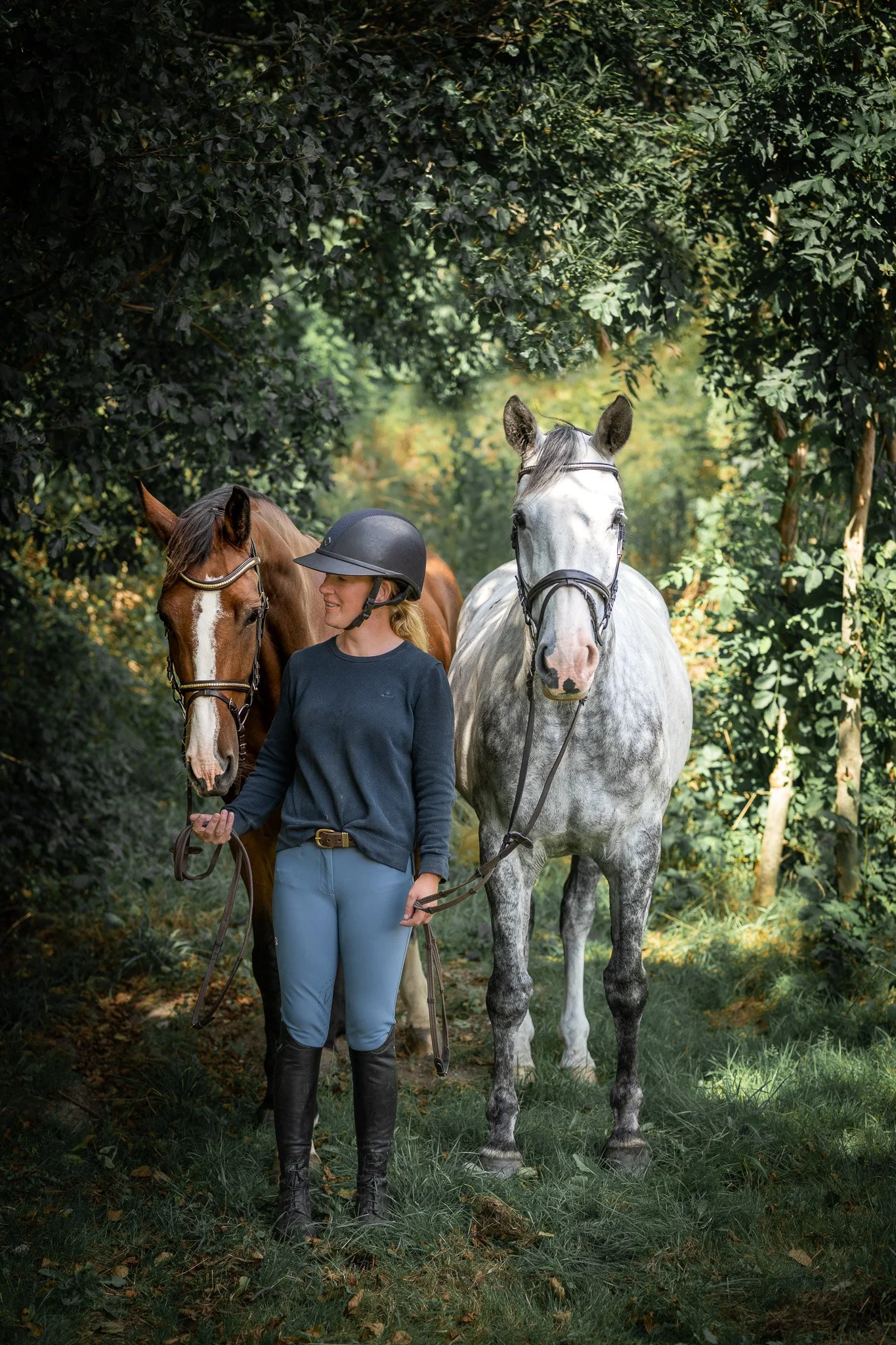 A woman wearing equestrian riding attire, including a helmet, standing in a forest with two horses, one brown and one gray, on a trail surrounded by dense green foliage.