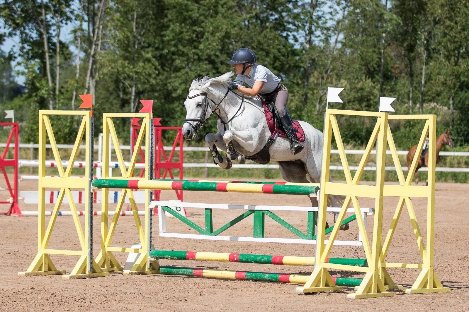 A person riding a white horse jumping over a colorful obstacle in an outdoor equestrian arena, with another horse visible in the background and trees surrounding the area.