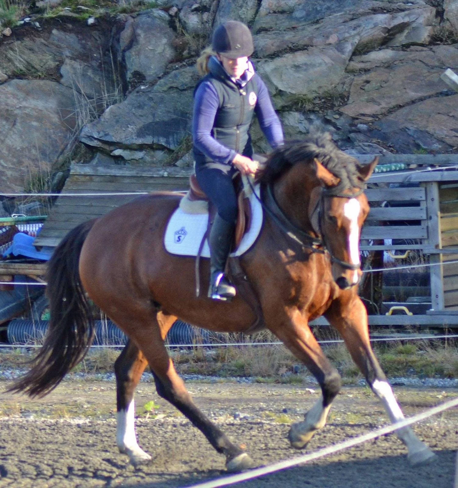 A woman riding a brown horse on a dirt track with rocks and wooden structures in the background.
