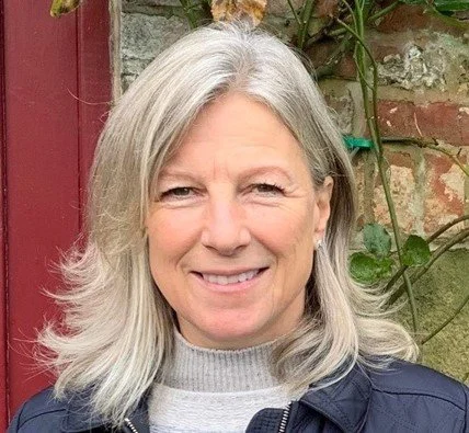 A woman with gray hair smiling outdoors in front of a brick wall and greenery.