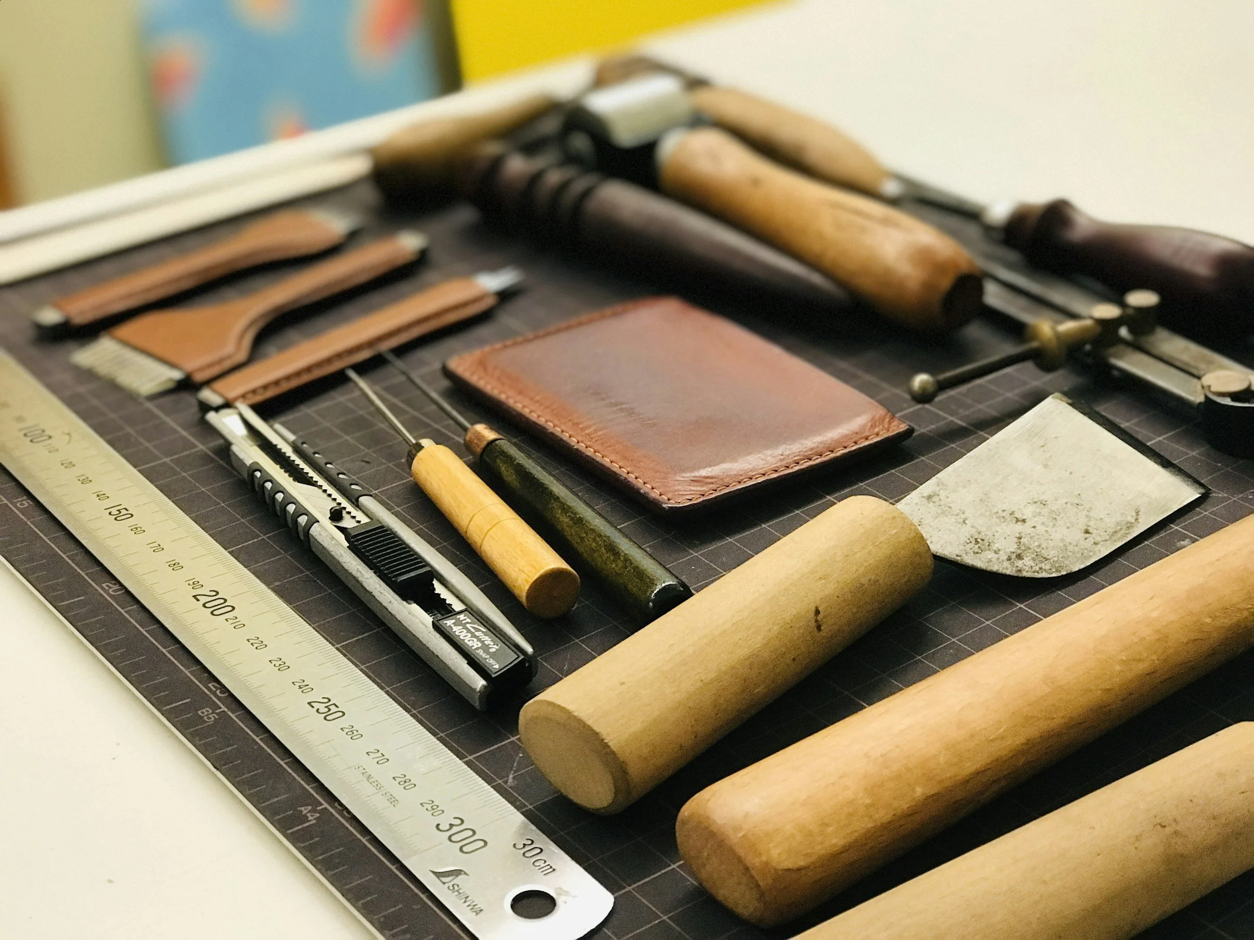 Leatherworking tools including a ruler, knives, punches, a stitching awl, a metal blade, and various wooden mallets, arranged on a cutting mat.
