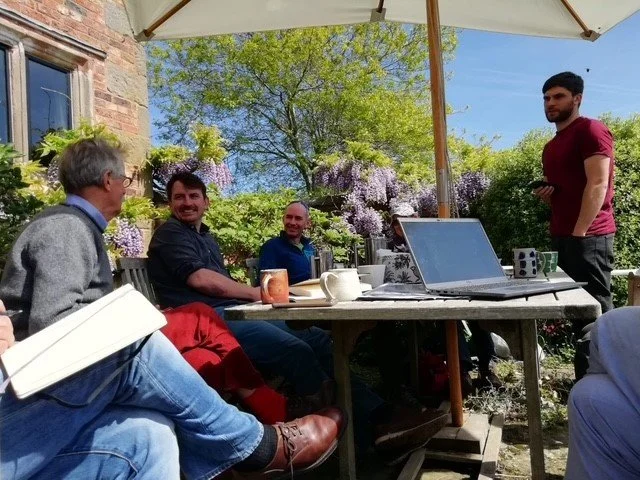 A group of people gathered around a table outdoors under a large umbrella, enjoying a sunny day with blooming trees and flowers in the background. They appear to be having a casual meeting or social gathering.