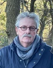 An older man with gray hair and glasses outdoors, wearing a dark blue jacket and a gray scarf, standing in front of a tree with green leaves.