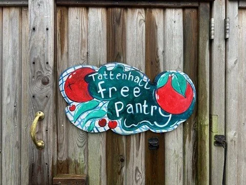 A colorful sign on a wooden fence reads 'Tattenhall Free Pantry' with illustrations of a tomato, cherry, and leaves.