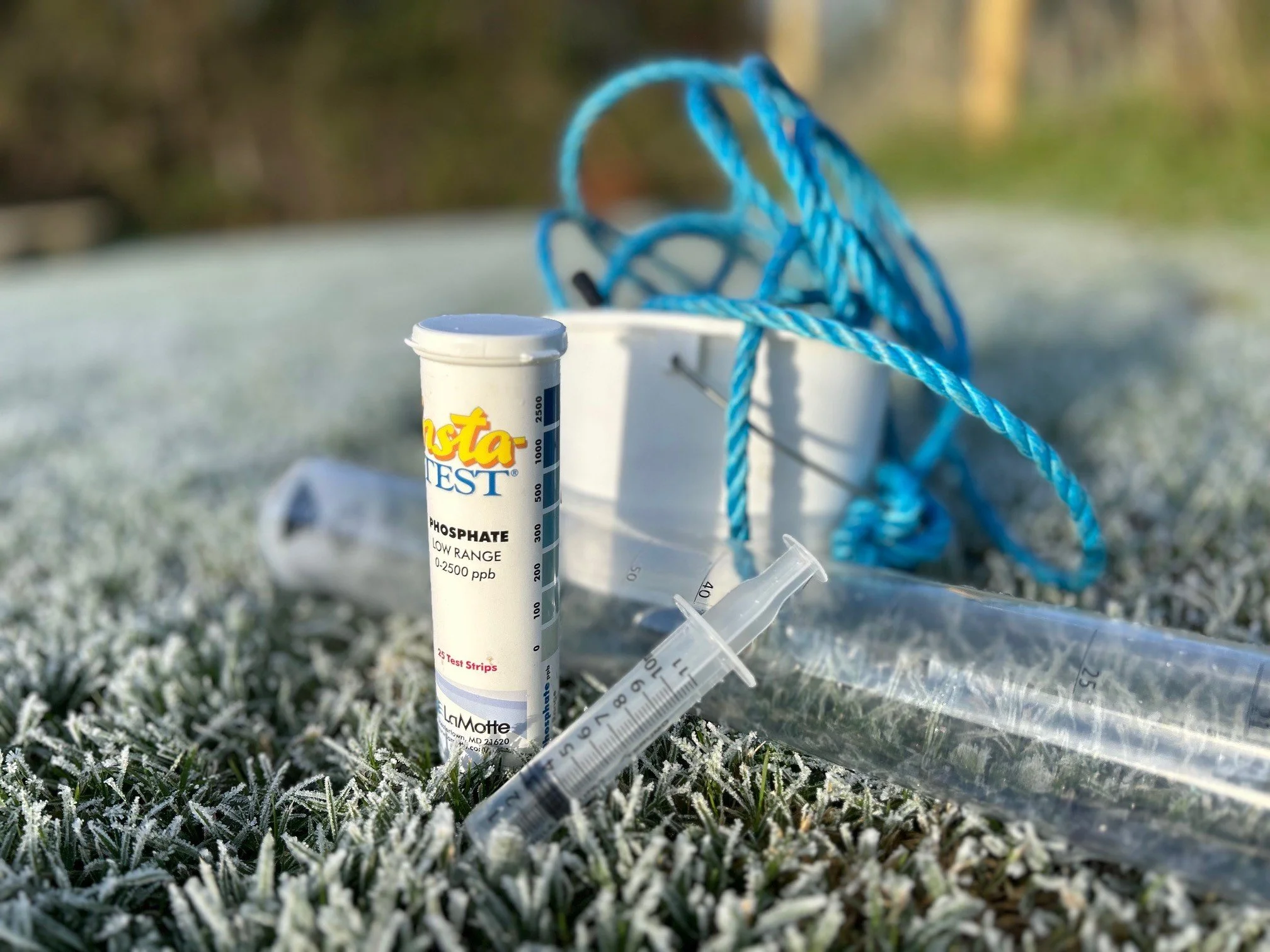 A close-up of a medical testing kit on frosty grass, including a container labeled 'Phosphate,' a syringe, a plastic tube, and blue string.