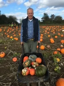 A man standing in a pumpkin patch with a basket of assorted pumpkins, smiling at the camera, with rows of pumpkins and a partly cloudy sky in the background.