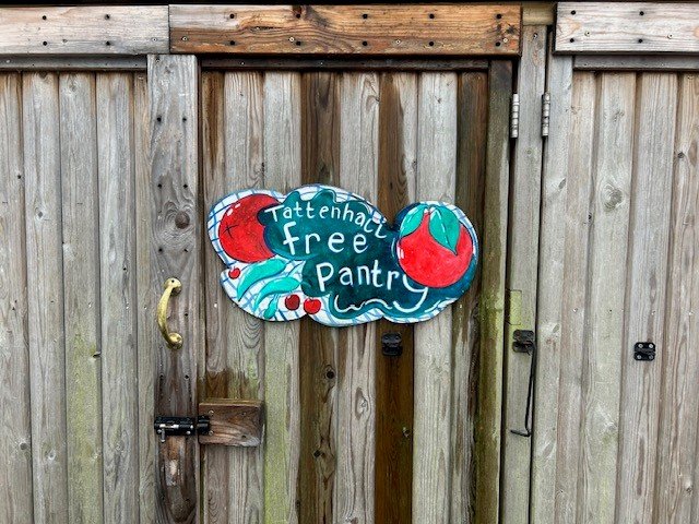 Colorful painted sign on wooden fence that reads "Tattenhaugh Free Pantry" with illustrations of tomatoes and other vegetables.
