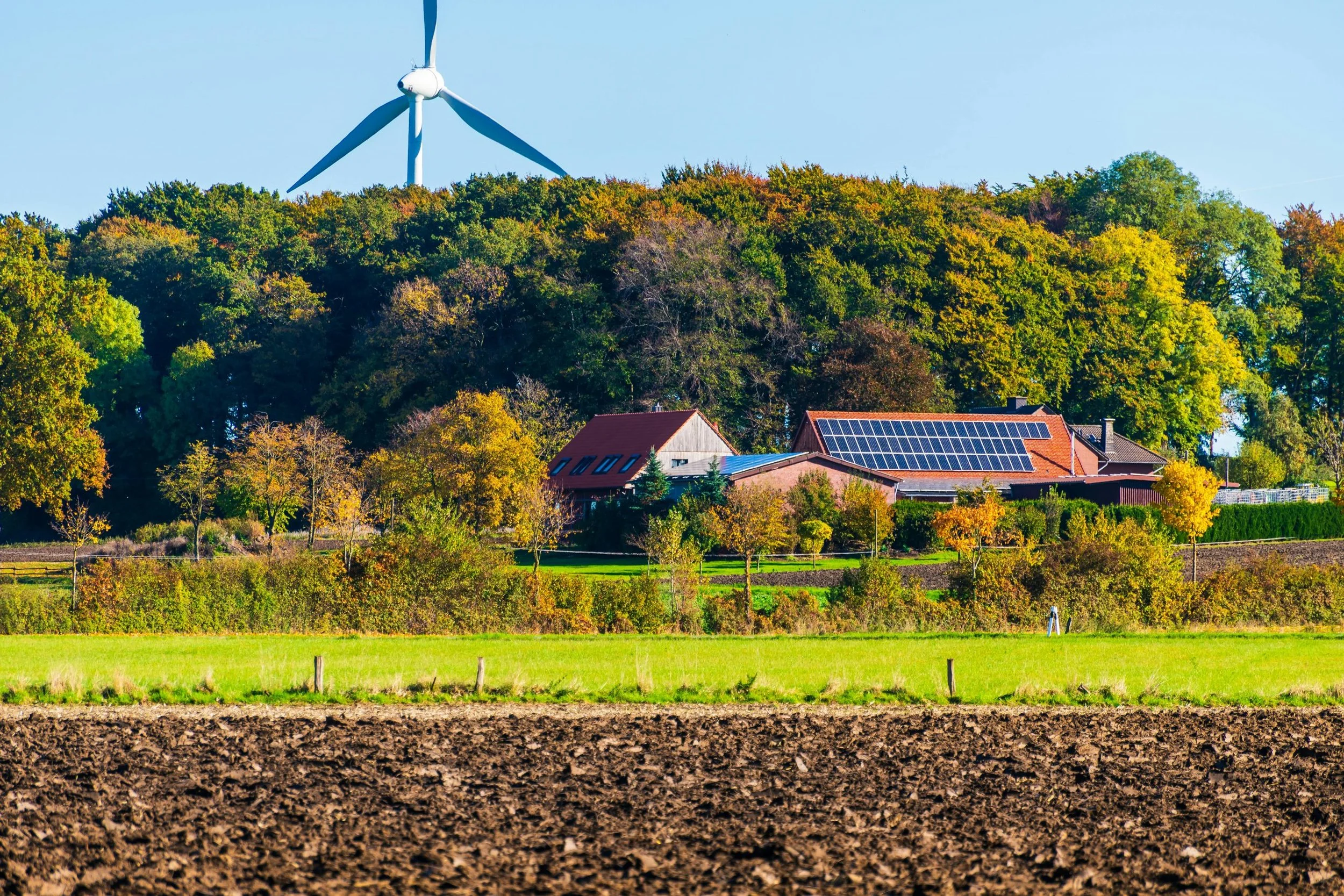 A scenic rural landscape with a wind turbine on a hill, trees with fall foliage, a house with solar panels on the roof, and a plowed field in the foreground.