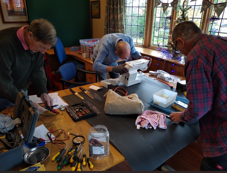 Three men working on electronic devices and sewing machines at a dark wood table in a room with large windows, Christmas decorations, and various tools and supplies scattered on the table.
