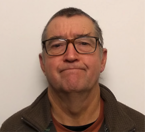 A middle-aged man with short hair, wearing glasses, a puzzled or serious expression, and a brown jacket against a plain white background.