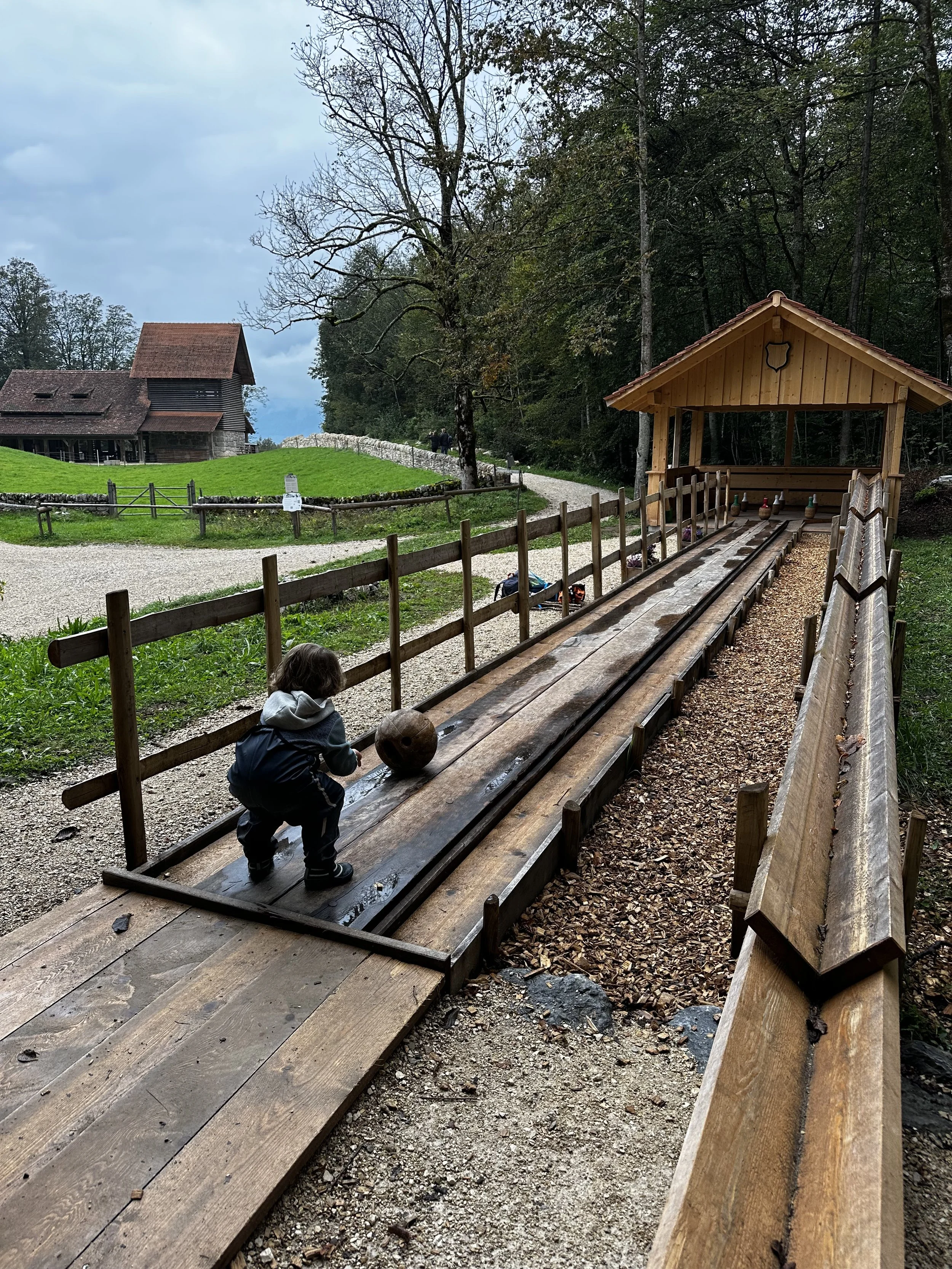 A child crouched down on a wooden alleyway at a rustic outdoor bowling game, with a large wooden ball in front of them, surrounded by a simple wooden fence, trees, and old buildings in the background.