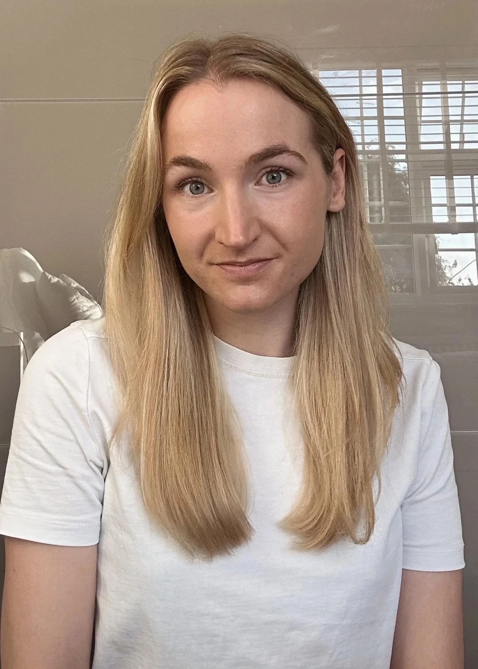 A young woman with long blonde hair wearing a white t-shirt, sitting indoors near a window with blinds, looking at the camera.