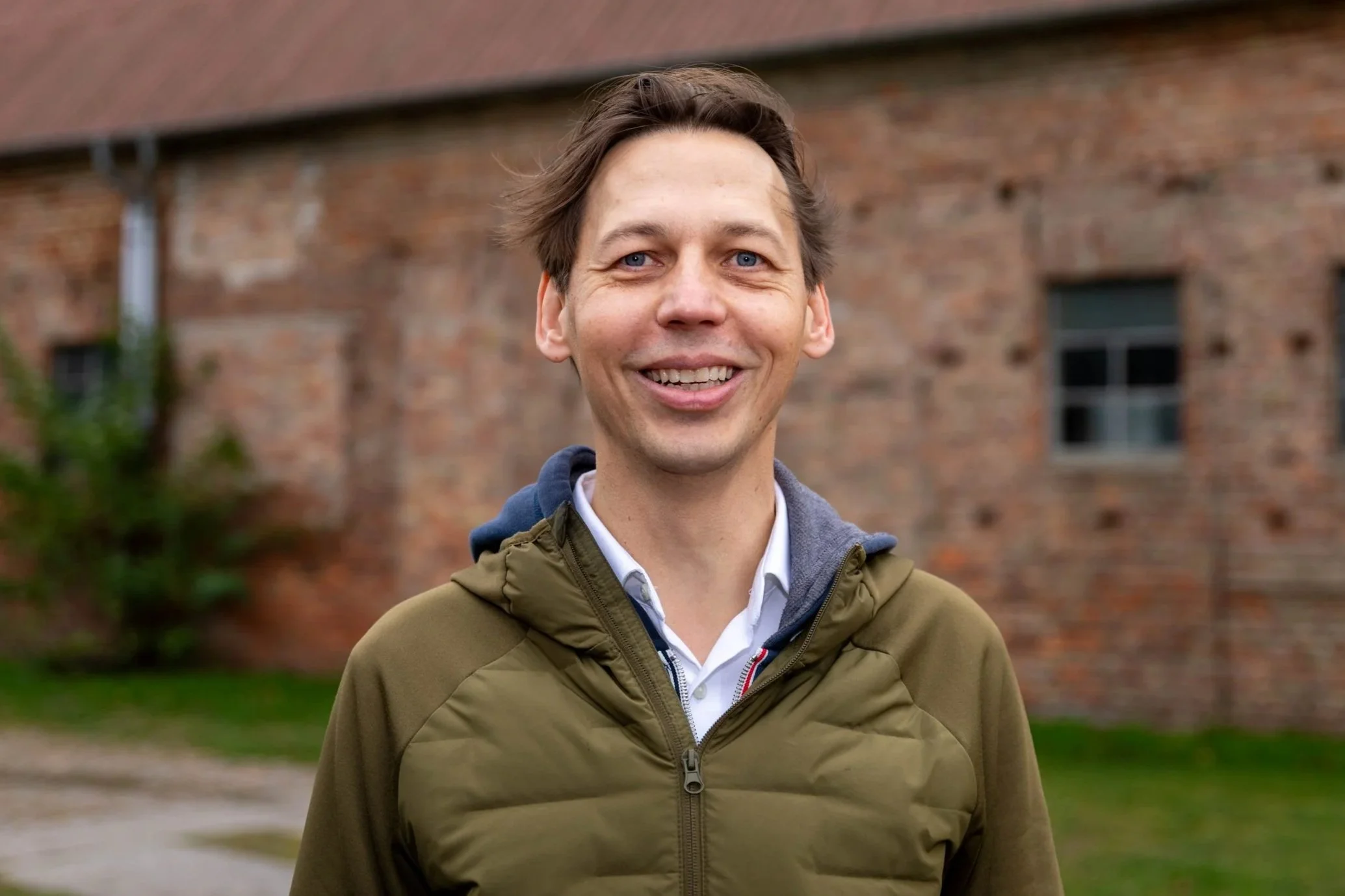 Jan-Henrik Hempel trägt eine olivgrüne Jacke und lächelt in die Kamera. Hinter ihm sieht man die Backsteinmauer eines Gebäudes.